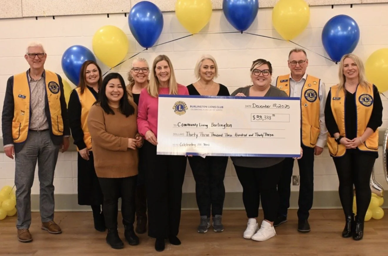Group of ten people standing indoors, holding a large check for $33,333 made out to Community Living Burlington, with yellow and blue balloons in the background.