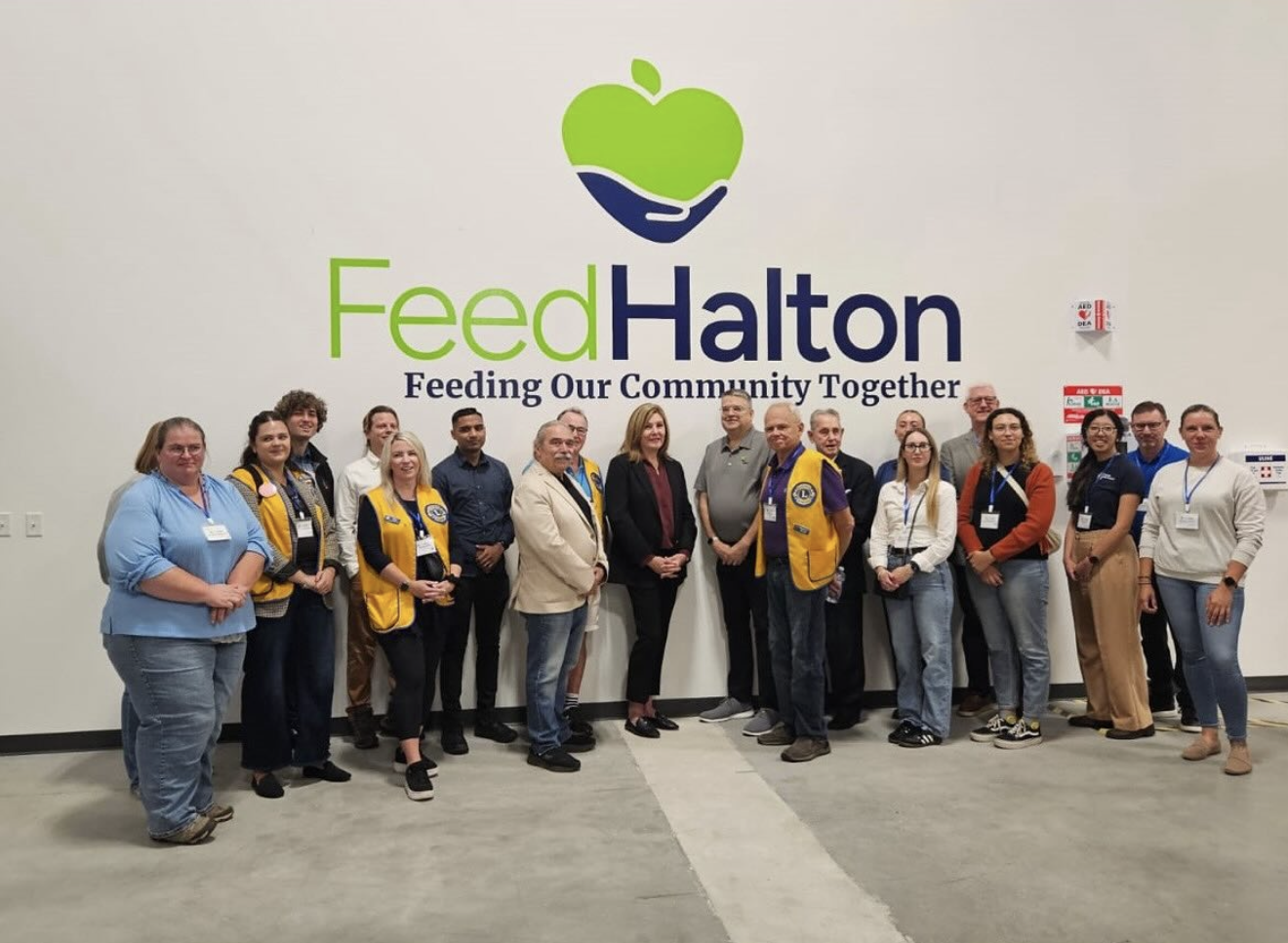 Group of diverse people standing in front of a wall with the Feed Halton logo and slogan, some wearing yellow vests, at a community event.