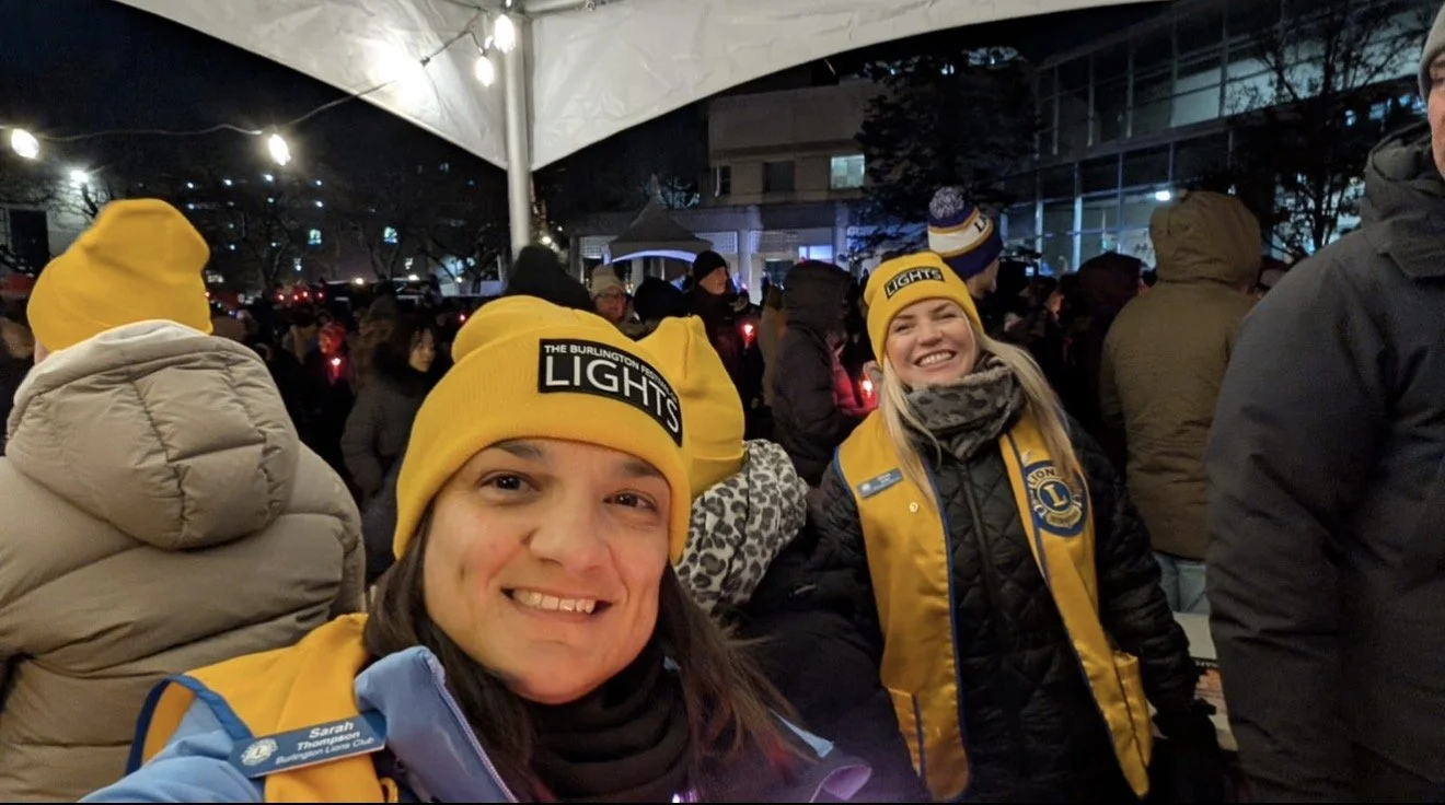 Two women wearing yellow knit hats and yellow vests with Lions Club logos, smiling at a nighttime outdoor event with a crowd and string lights.
