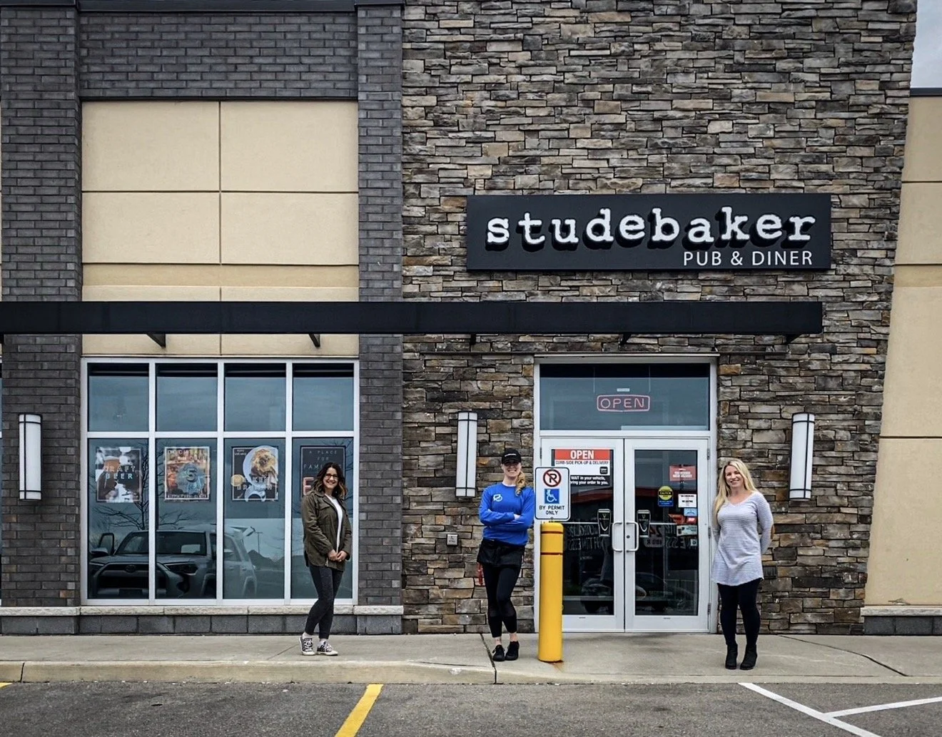 Three women standing in front of the Studebaker pub and diner, which has a brick exterior and large windows. One woman is on the left near the window, the second in the center wearing a blue jacket and black cap, and the third on the right is smiling in a white striped top. There are signs on the door including an 'Open' sign and parking lot markings in the foreground.
