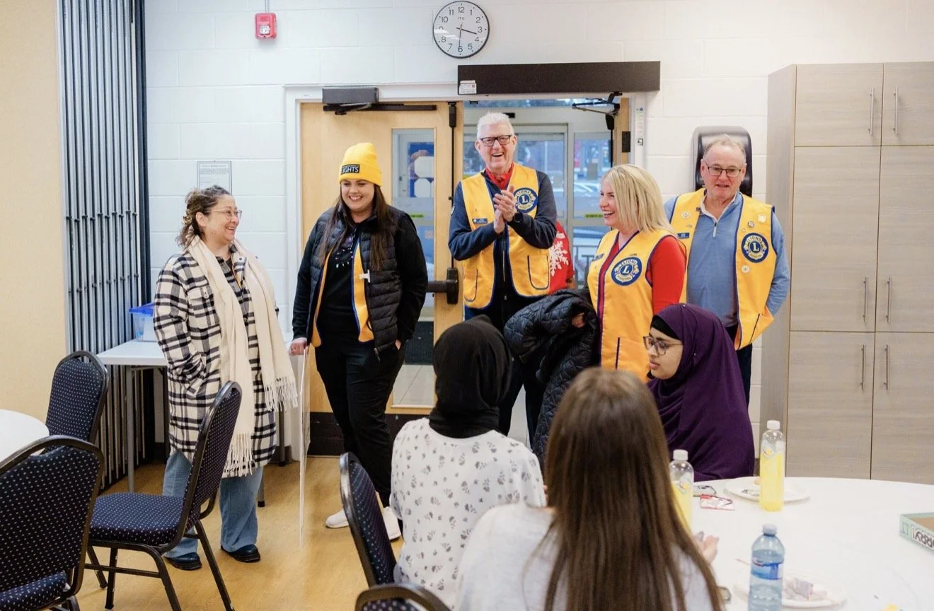 Group of people in a community center, with some seated at a round table and others standing, wearing yellow vests with a Lions Club logo, engaged in a lively discussion or presentation.