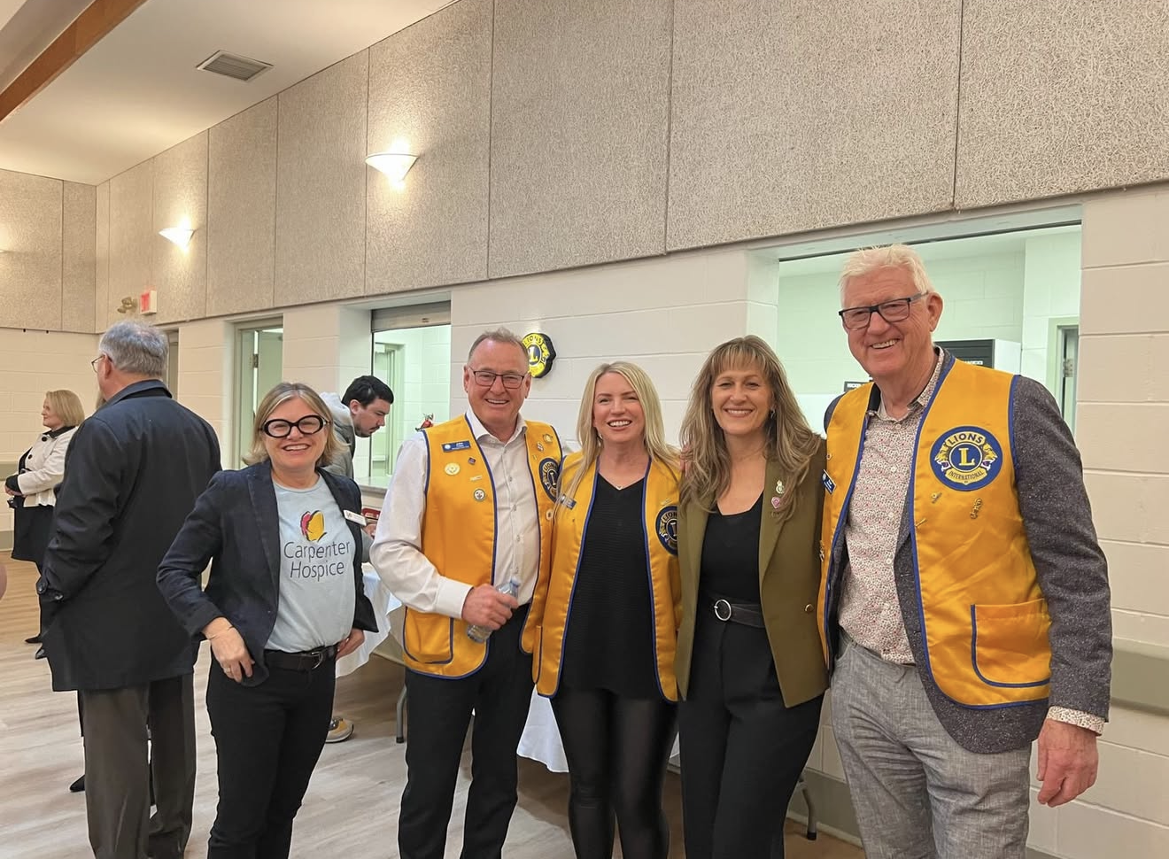 A group of five people smiling for a photo at a social event. Four individuals are wearing vests with Lions Club logos, and one woman is wearing a t-shirt with Carpenters Hospice logo. They are standing indoors in a room with beige walls and soft lighting.