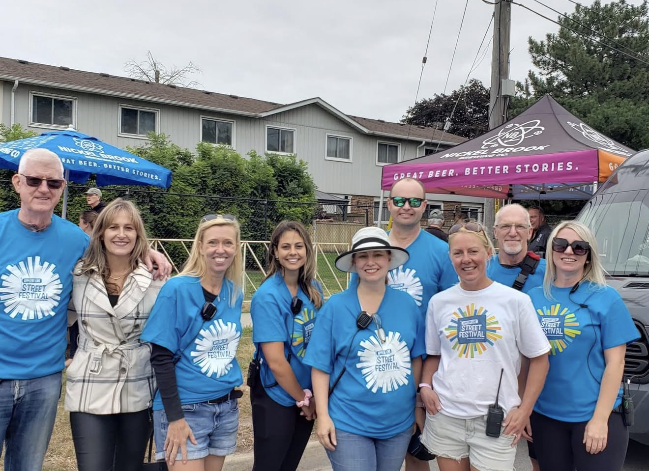 Group of people wearing blue and white shirts at an outdoor street festival, with tents and a residential building in background.