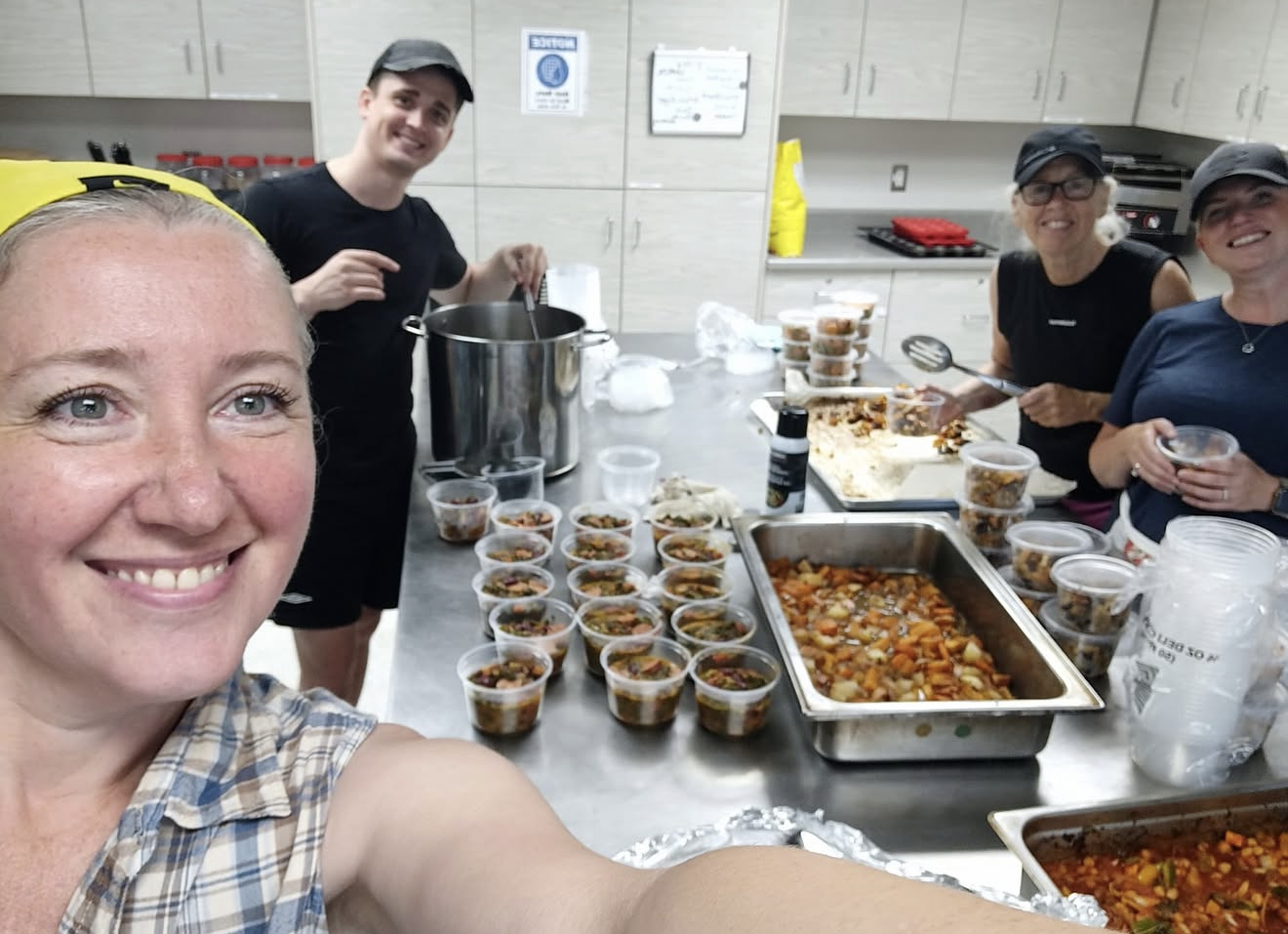 Four women and one man in a kitchen preparing and serving food, with several small cups filled with a vegetable stew on the table.