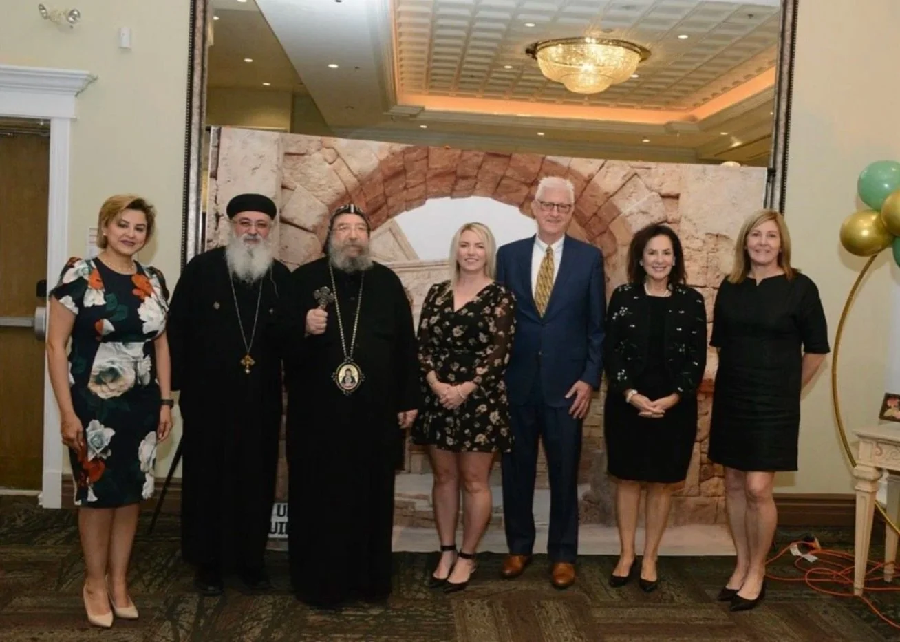 Group of seven people dressed in formal attire standing in front of a backdrop resembling a stone archway in an indoor event space, with balloons on the right side.