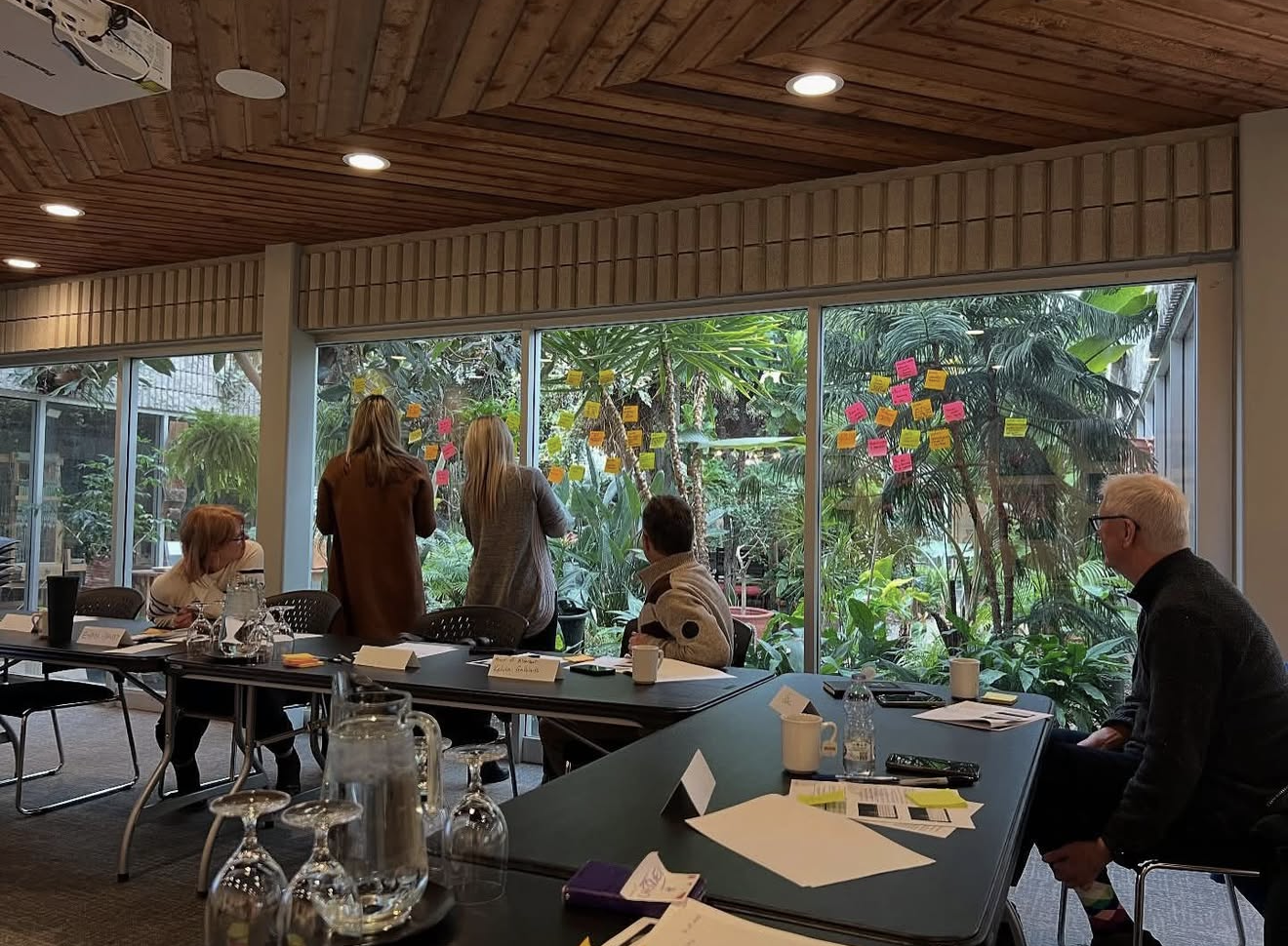 People in a conference room observing and possibly discussing notes on sticky notes on a large window overlooking a garden.