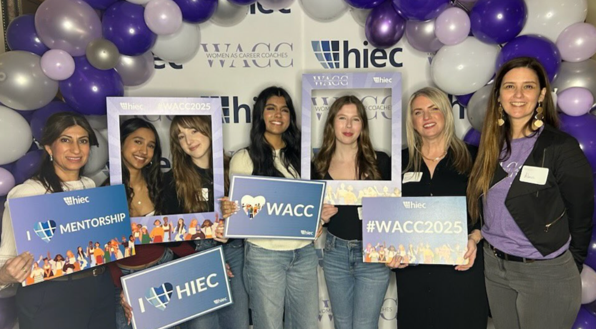 Group of six women at a WACC organized event, holding signs about mentorship, #WACC2025, and love for HIEC. Background decorated with purple, white, and silver balloons, and a banner with the WACC and HIEC logos.
