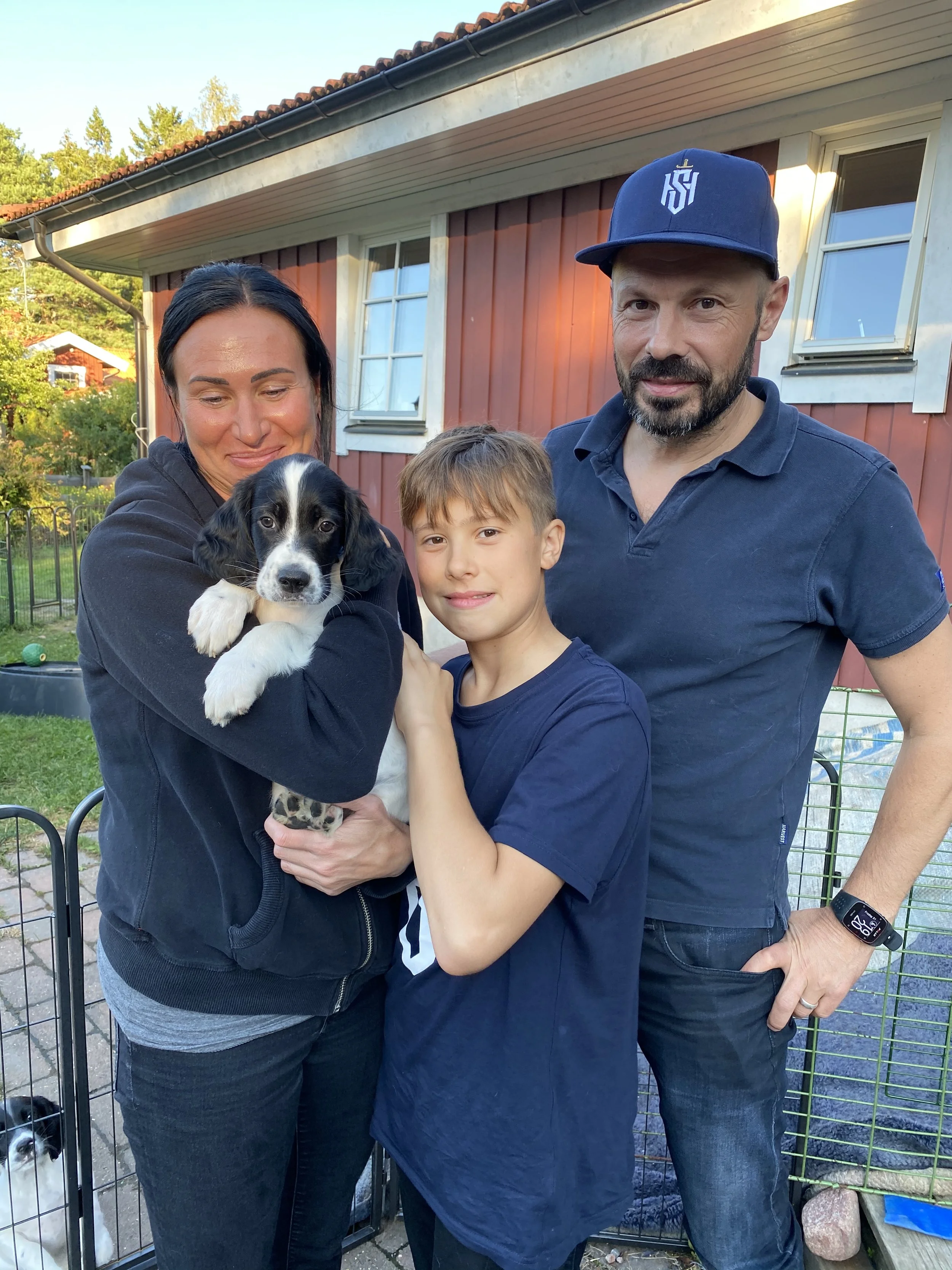 En familj med två vuxna och en pojke, alla i casualkläder, står utomhus framför ett rött hus med vita fönster och håller en svart och vit valp.