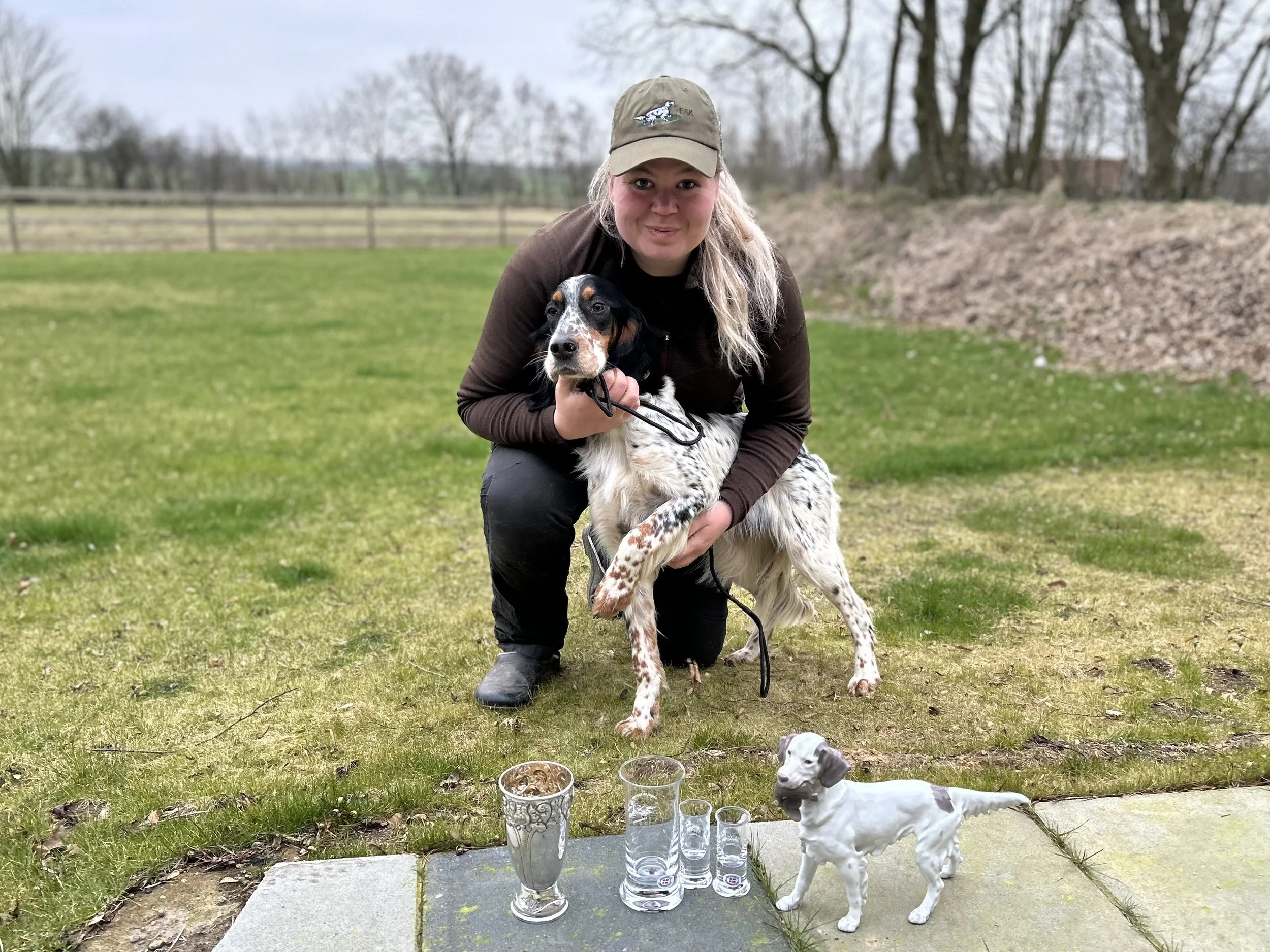 En kvinna i mössa ler och håller i en hund med svart, vitt och brunt päls på en gräsmatta, andra hundar ligger bredvid henne, och det finns drycker i små glas på en stenplatta framför.