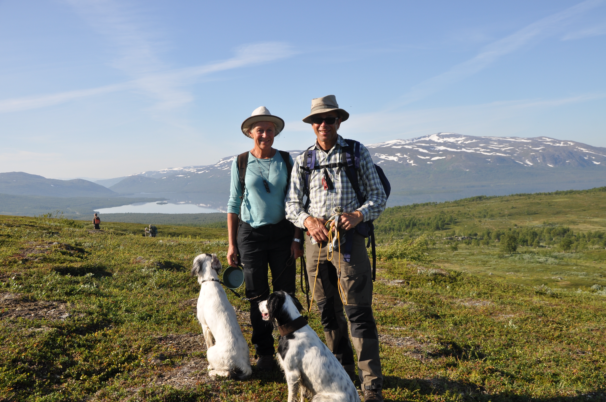 Två personer, en man och en kvinna, står på en grön äng med en hund, med snötäckta berg i bakgrunden och en klar blå himmel.