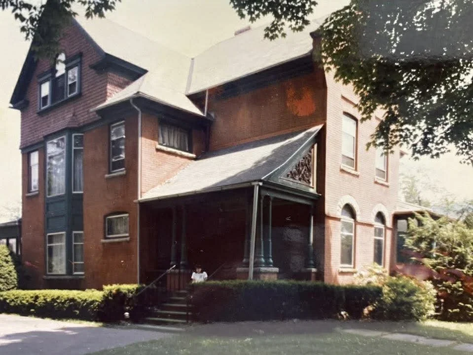 Circa 1980s - Jeanne McCloskey sits on the front porch. The color scheme of the exterior trim - Tuscan red shingles and dark green columns - was on the house at least from the 1950s and is thought to have been original to the 1890s.