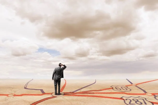 A person standing at a desert intersection of painted highway lines and looking up at the cloudy sky.