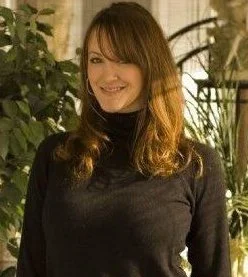 A woman with shoulder-length brown hair smiling, wearing a black turtleneck, surrounded by indoor plants.