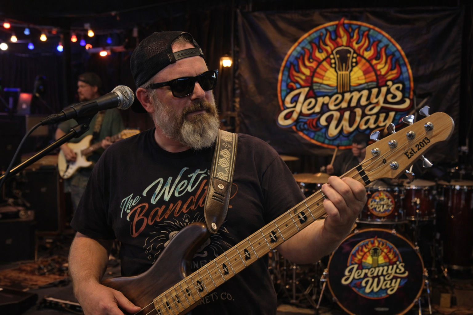 A bearded man wearing sunglasses, a black baseball cap backwards, and a black T-shirt with 'The Wet Bands' on it, playing a bass guitar. In the background, there is a drum set and a banner with a colorful logo reading 'Jeremy's Way'. A guitarist is also seen in the background.