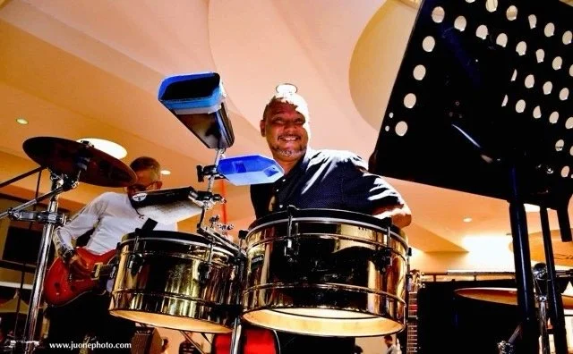 A smiling man playing steel drums in a band, with another musician playing guitar in the background, in an indoor setting.