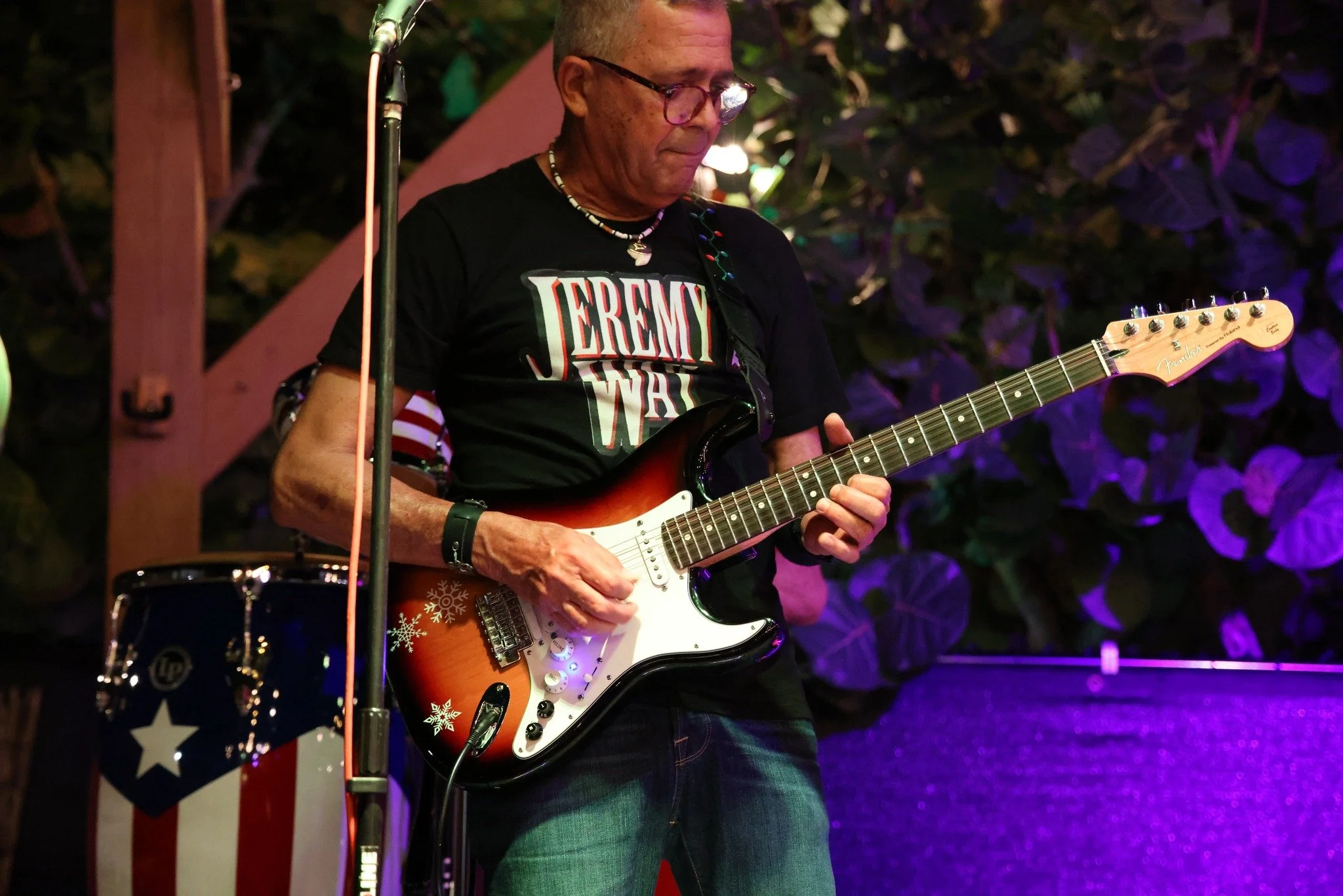 A man playing an electric guitar on stage, wearing a black T-shirt with the words 'JEREMY WASHINGTON' and glasses, with music equipment and purple lighting in the background.