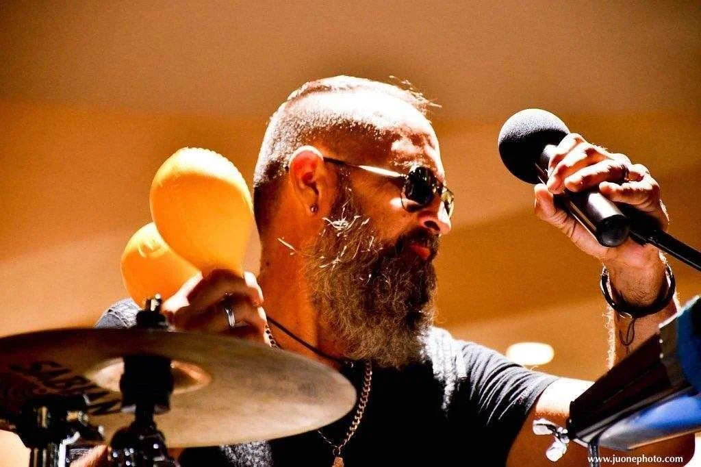 A man with a beard and sunglasses holding a pair of maracas in one hand and a microphone in the other, with a drum set in front of him.