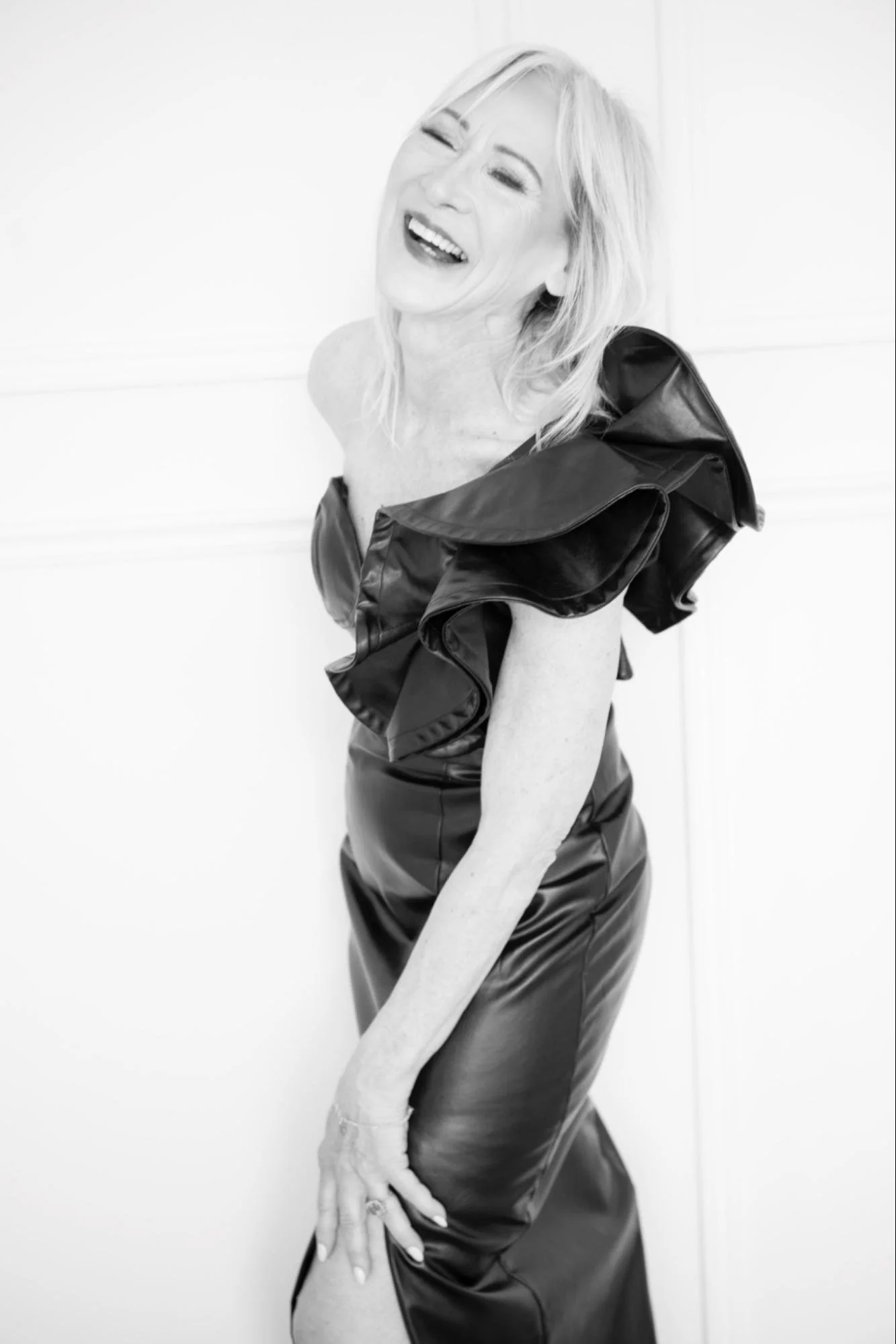A woman with blonde hair laughing, wearing a black shiny dress with ruffled shoulders, standing against a plain white background.