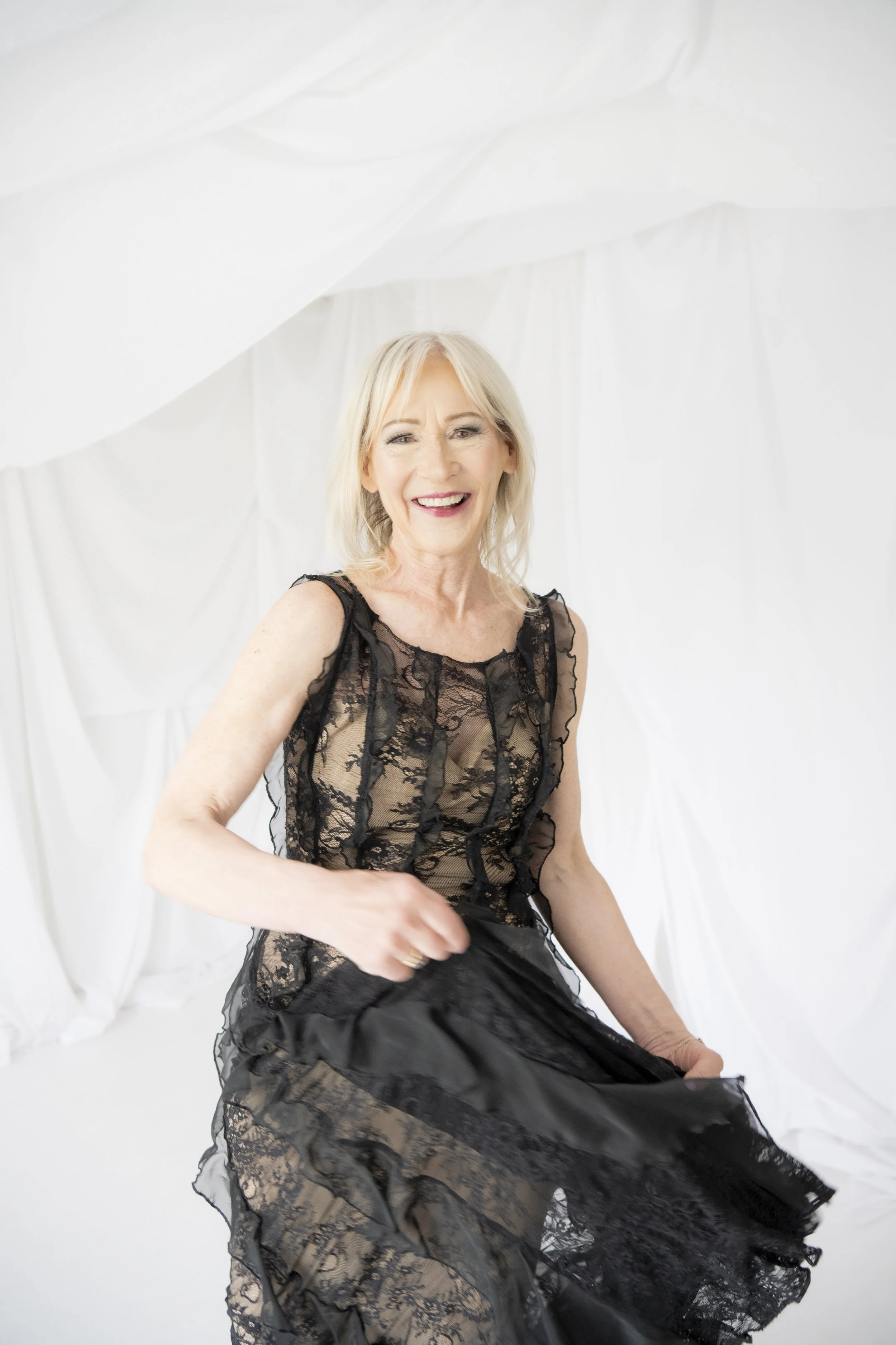 A smiling woman in a black lace dress standing against a white backdrop, holding the hem of her dress.