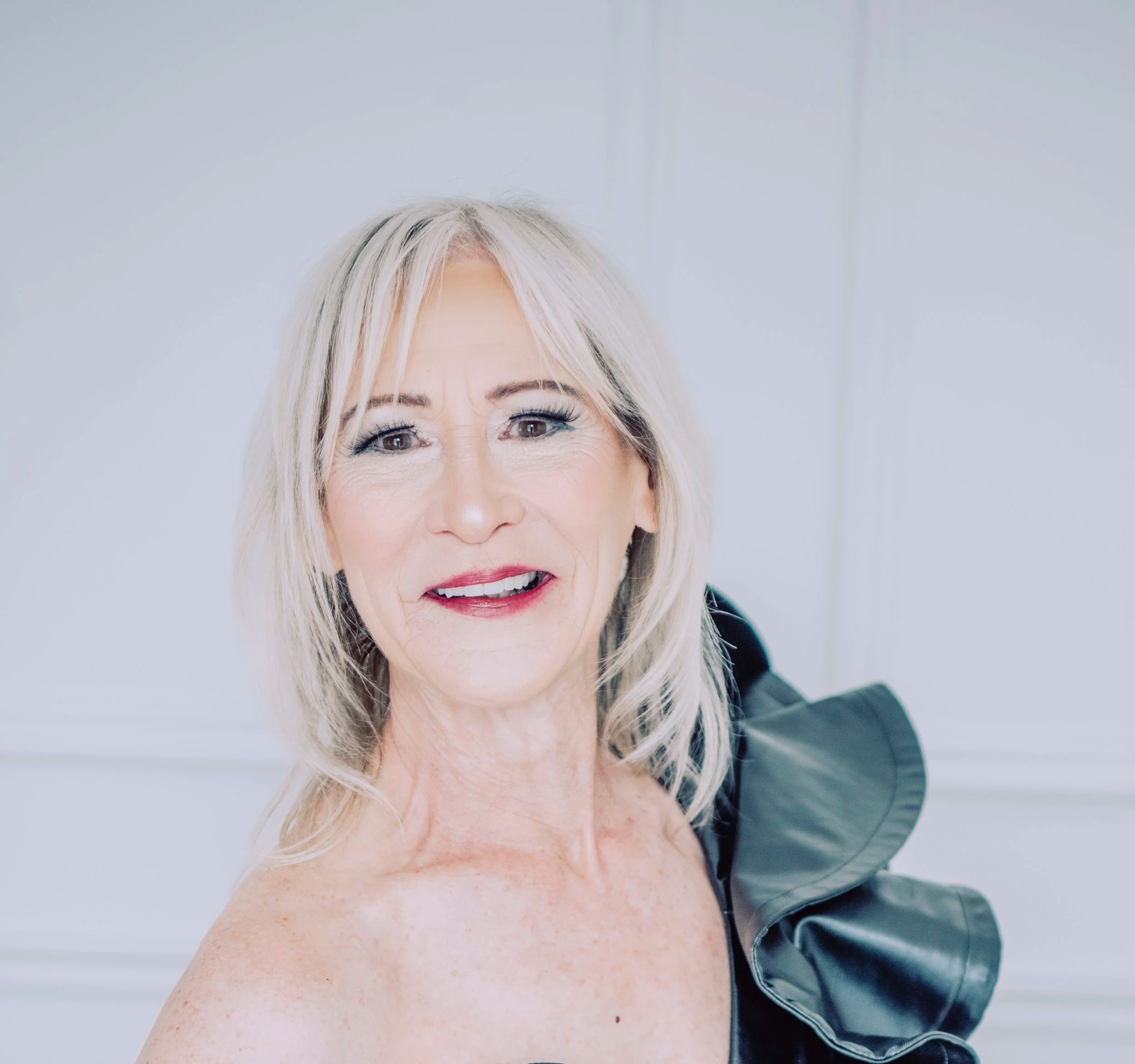 Valerie Maxemniuk  smiling at the camera, wearing a black garment with ruffle details on her shoulder, in a light-colored room.