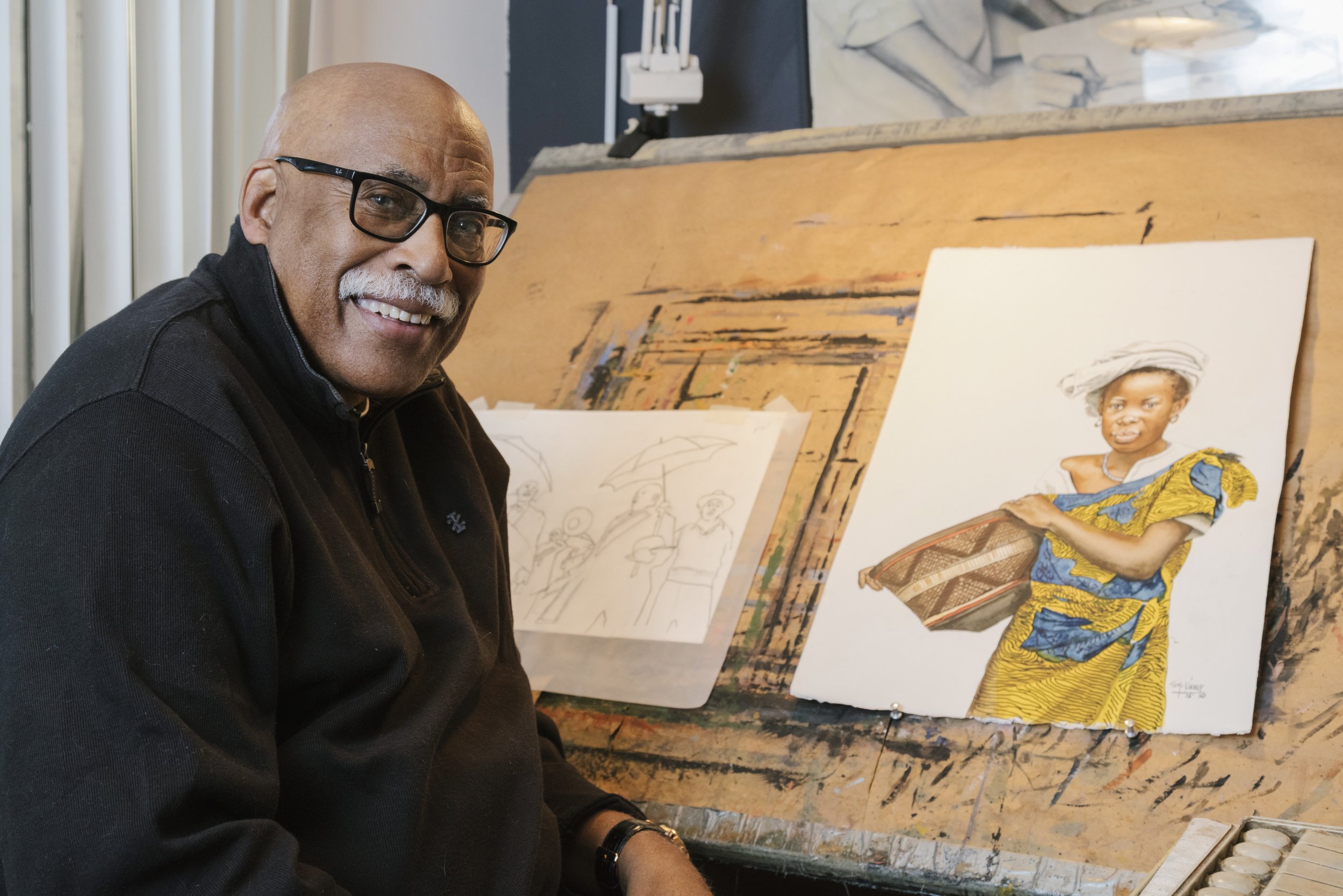 Watercolorist Tom McKinney working on a colorful painting of a woman in traditional African clothing and headwrap, sitting at an easel in an art studio.