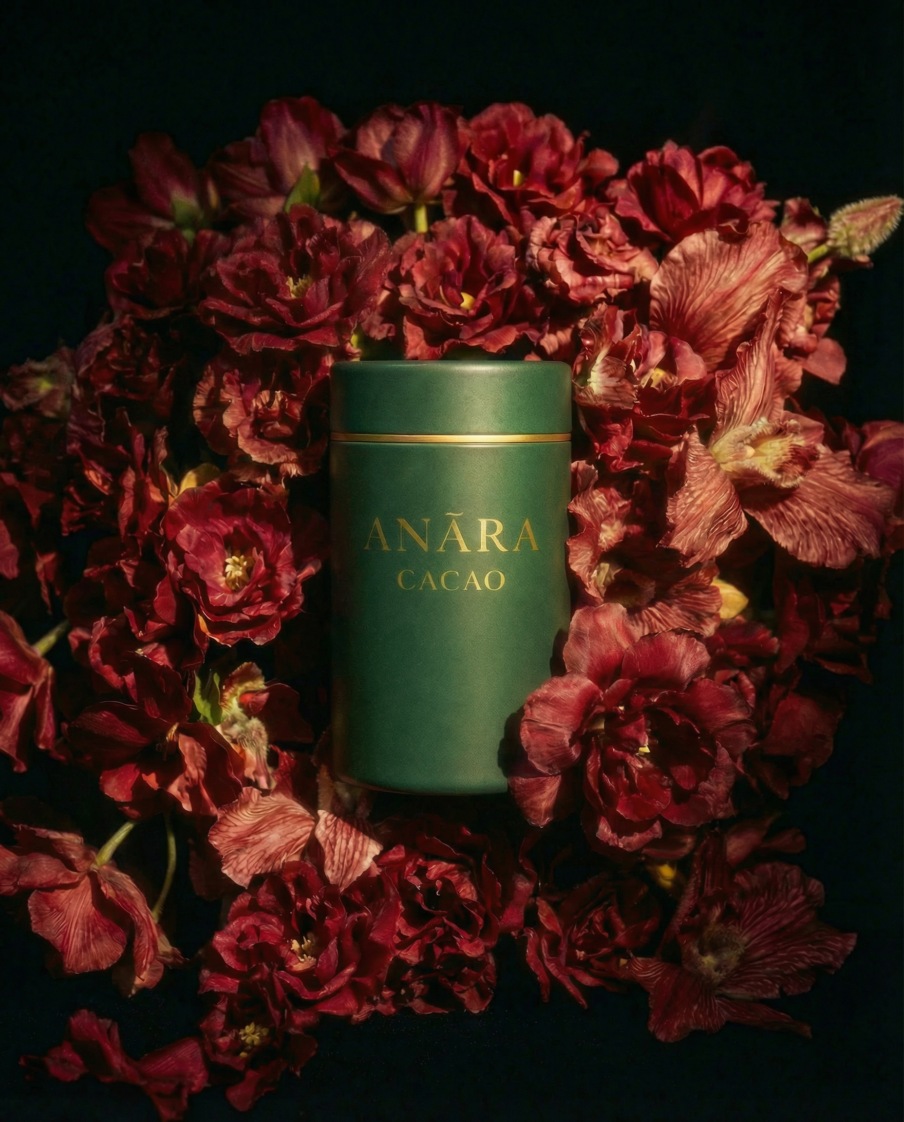 A green cylindrical container with gold lettering that reads 'ANARA CACAO' surrounded by red/burgundy flowers with some green leaves.