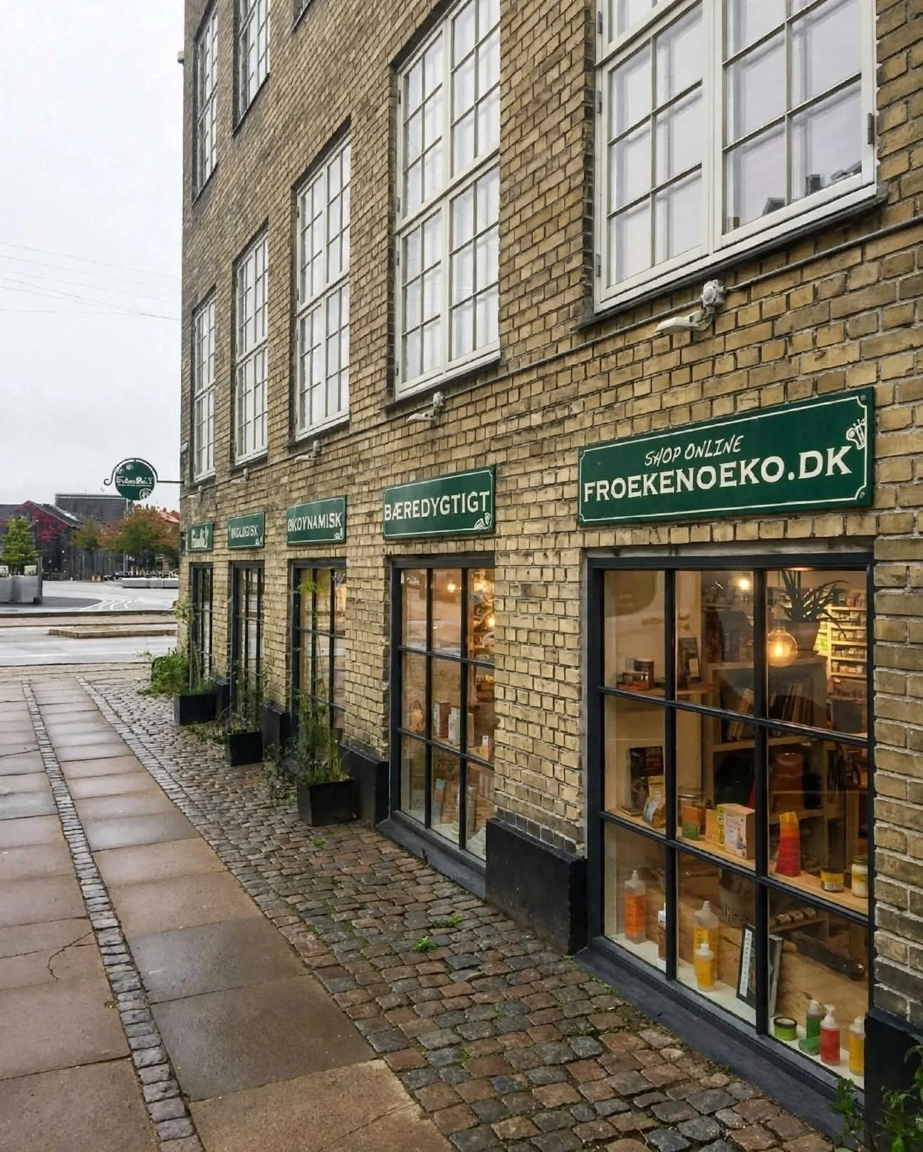 View of a brick building with large windows and green shop signs reading 'Shop Online Froekenoeko.dk', 'Bæredygtigt', 'Body Mask', and others. There are potted plants along the sidewalk and street in the background.