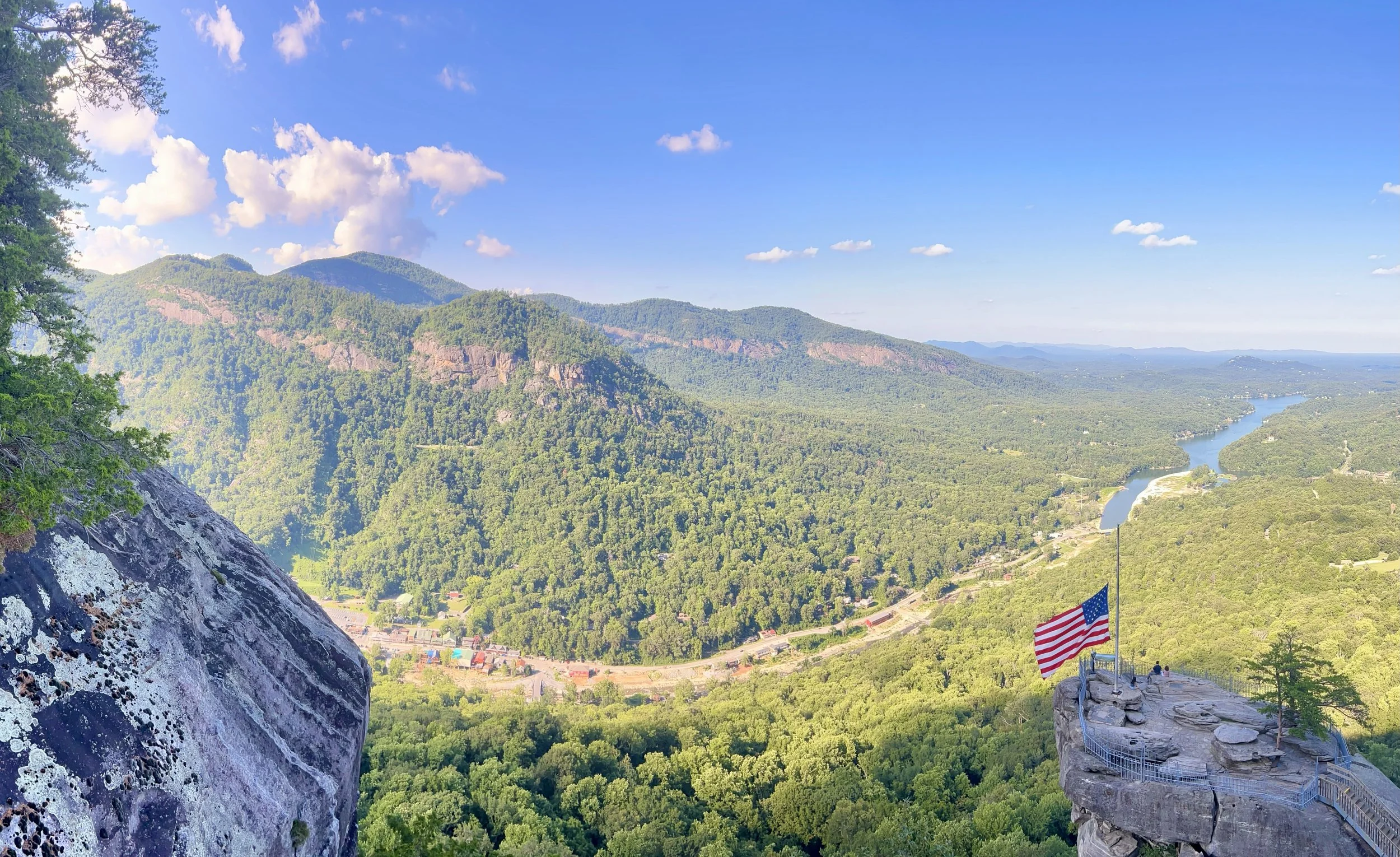 A scenic mountain landscape with lush green forested hills, a river winding through the valley, and a blue sky with scattered clouds. An American flag is visible on a rocky outcrop with a viewing platform.