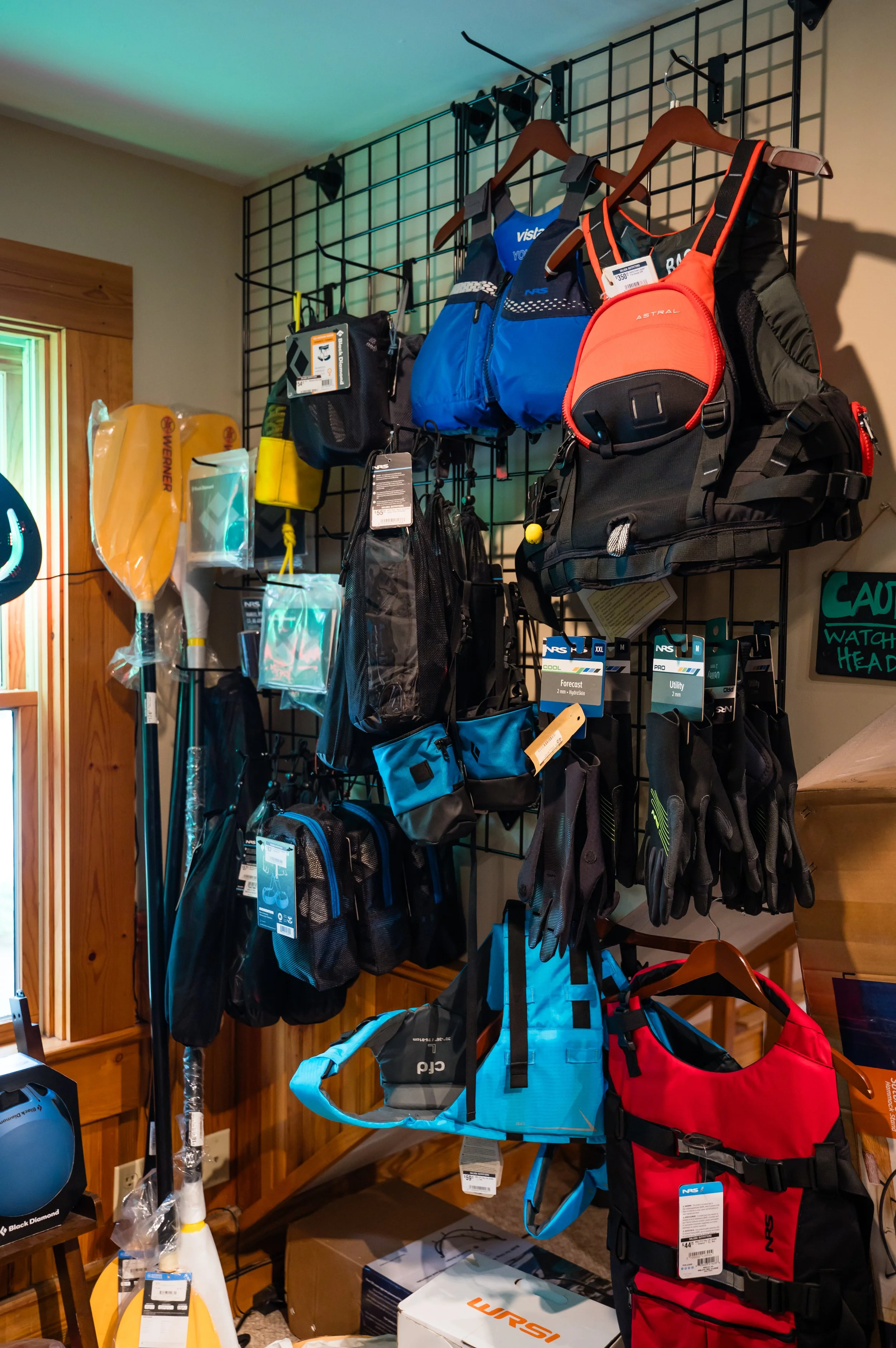 Display of outdoor gear including backpacks, hydration packs, gloves, and paddles on a metal grid wall in a store.