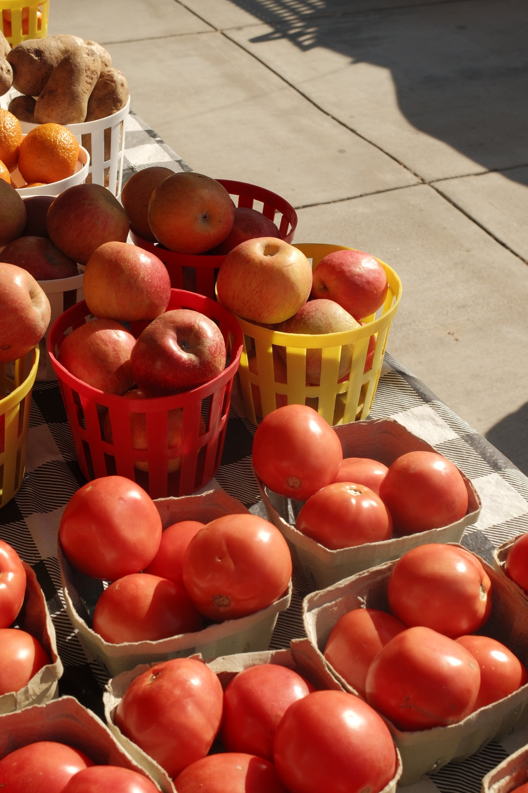 Baskets of fresh apples, tomatoes, oranges, and potatoes arranged on a table outdoors.