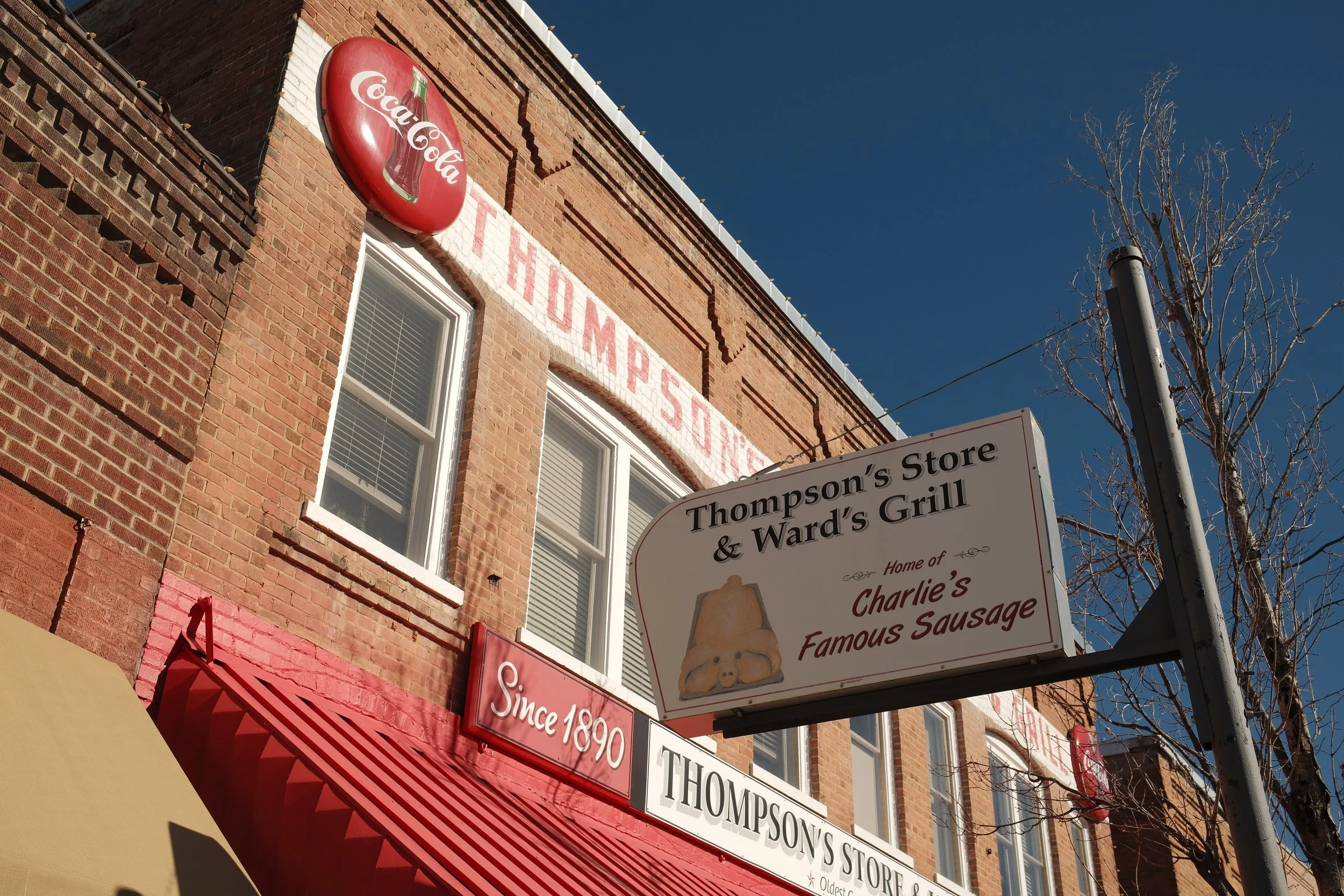 The image shows the exterior of a brick building with a vintage Coca-Cola sign and a sign for Thompsons Store. There is also a street sign that mentions Charlie's Famous Sausage and Ward's Grill. The sky is clear and blue, and there is a leafless tree beside the building.