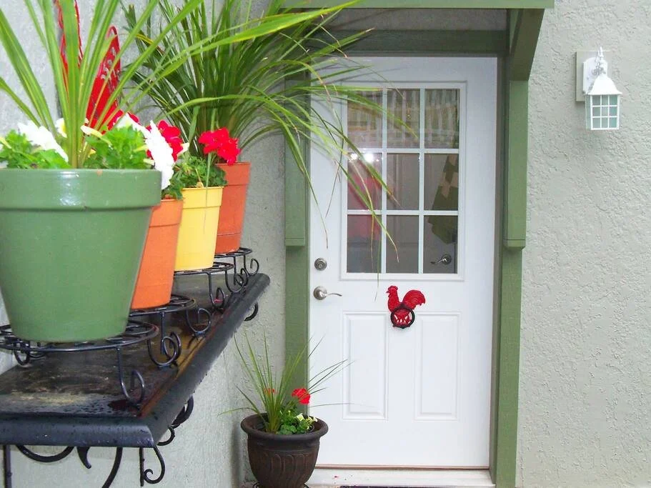 View of a front door decorated with a red rooster ornament, surrounded by potted plants, with a shelf of colorful pots on the left and a wall-mounted light fixture on the right.