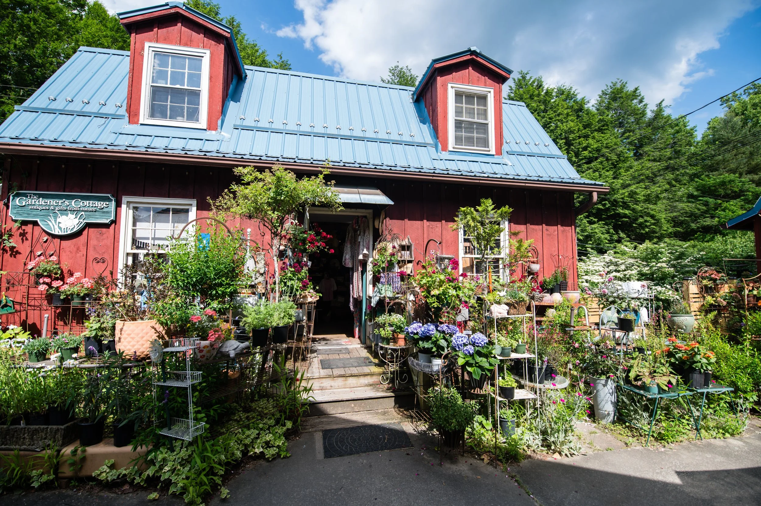 A red garden cottage with a blue metal roof and two dormer windows, surrounded by a lush garden with various flowers and plants, with a sign that reads 'The Gardener's Cottage'.