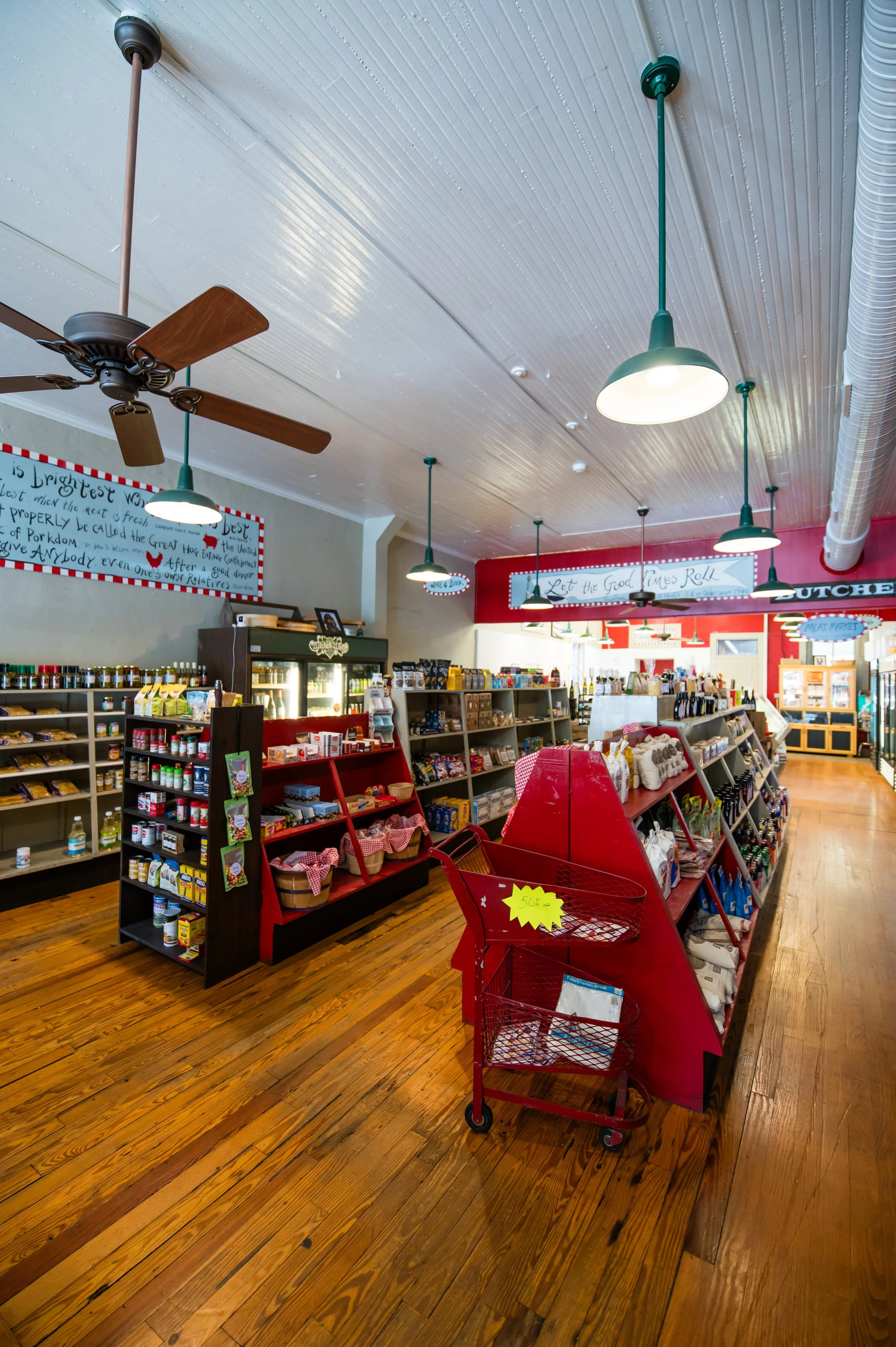 Interior of a grocery or retail store with shelves of products, Christmas-themed displays, a red shopping cart, and ceiling fans and lighting.