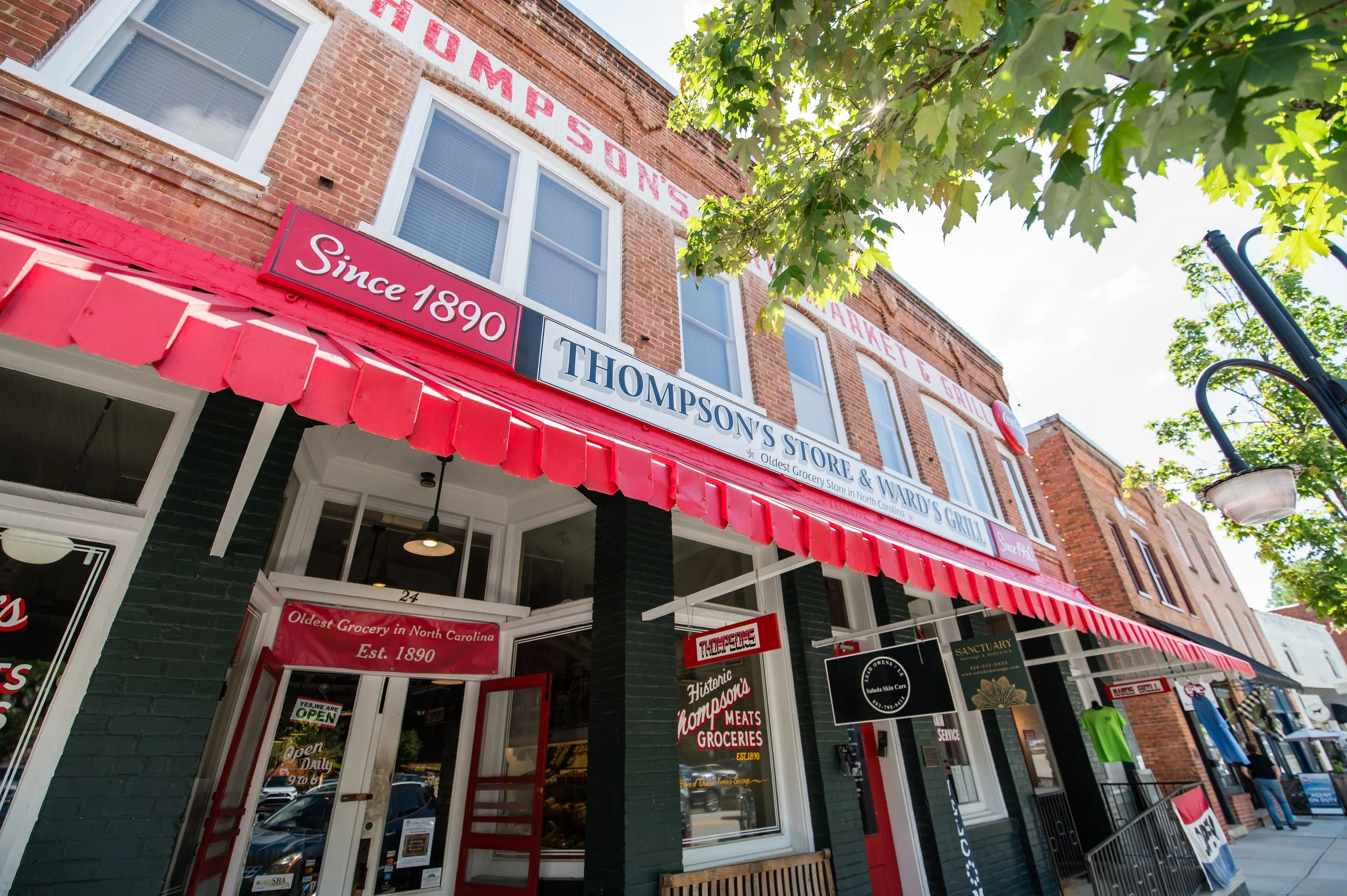 The exterior of Thompson's Store & Ward's Grocery on a sunny day, with a red awning and signs indicating it has been in operation since 1890 and is the oldest grocery in North Carolina. The building is brick with windows and greenery from nearby tree