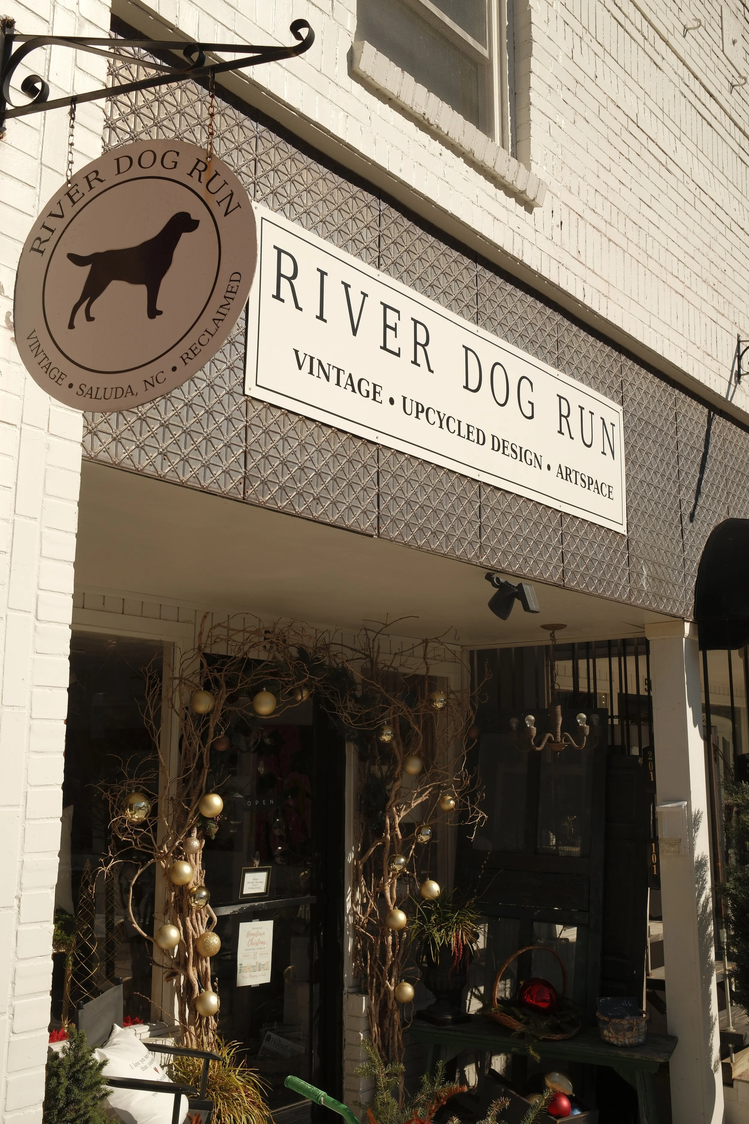 Storefront decorated for Christmas with a sign reading "River Dog Run," featuring a black silhouette of a dog. The display includes Christmas ornaments hanging from small trees in the window.