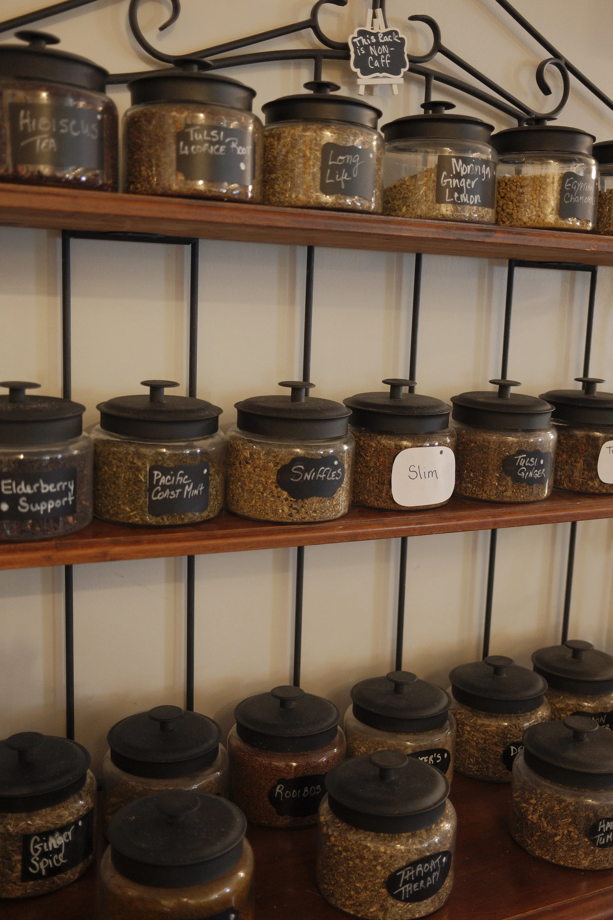 A wooden shelf displaying glass jars filled with various herbs and spices, each labeled with handwritten black chalkboard labels, with some labels reading 'Elderberry Support,' 'Pacific Coast Mint,' 'Snuffles,' and 'Ginger Spice.' There is also a sma