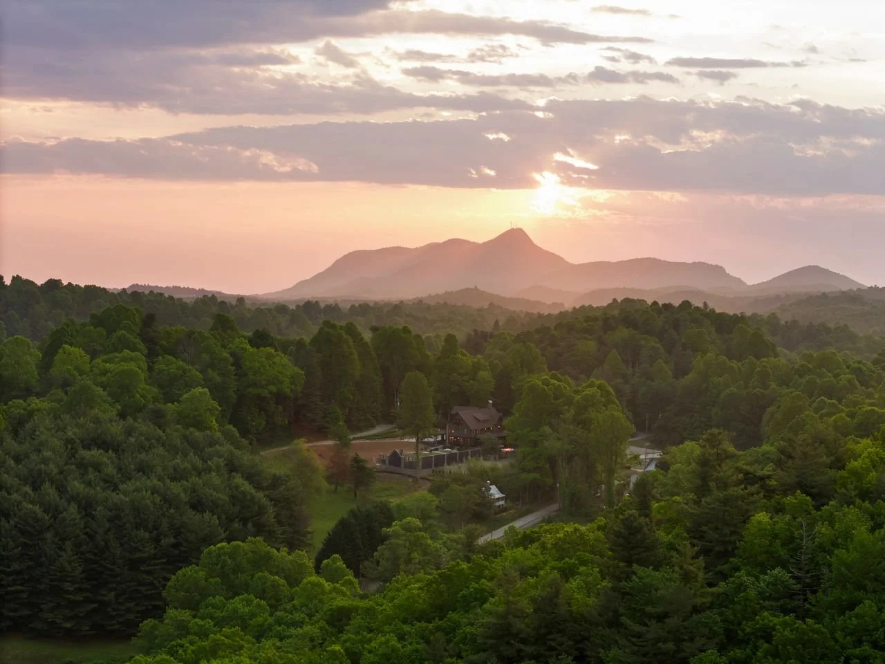 Scenic view of a lush green forest with a house and winding road in the foreground, mountains in the distance, and the sun setting behind clouds in the sky.