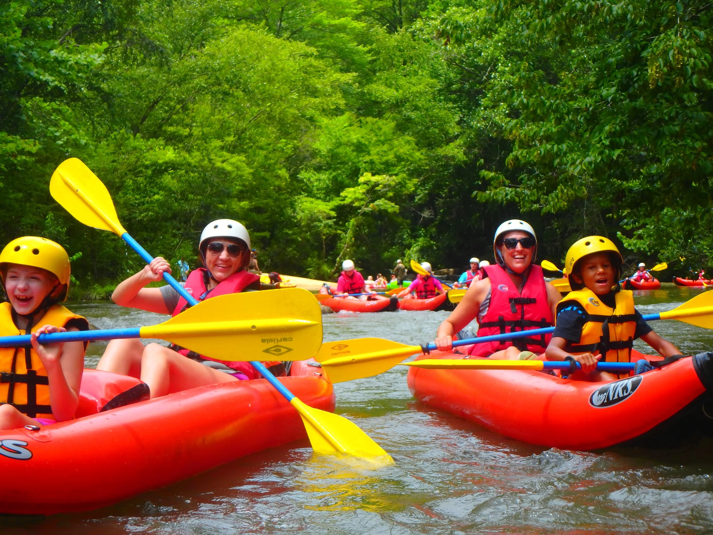 Group of smiling children and adults in yellow helmets and red life jackets kayaking on a river surrounded by lush green trees.
