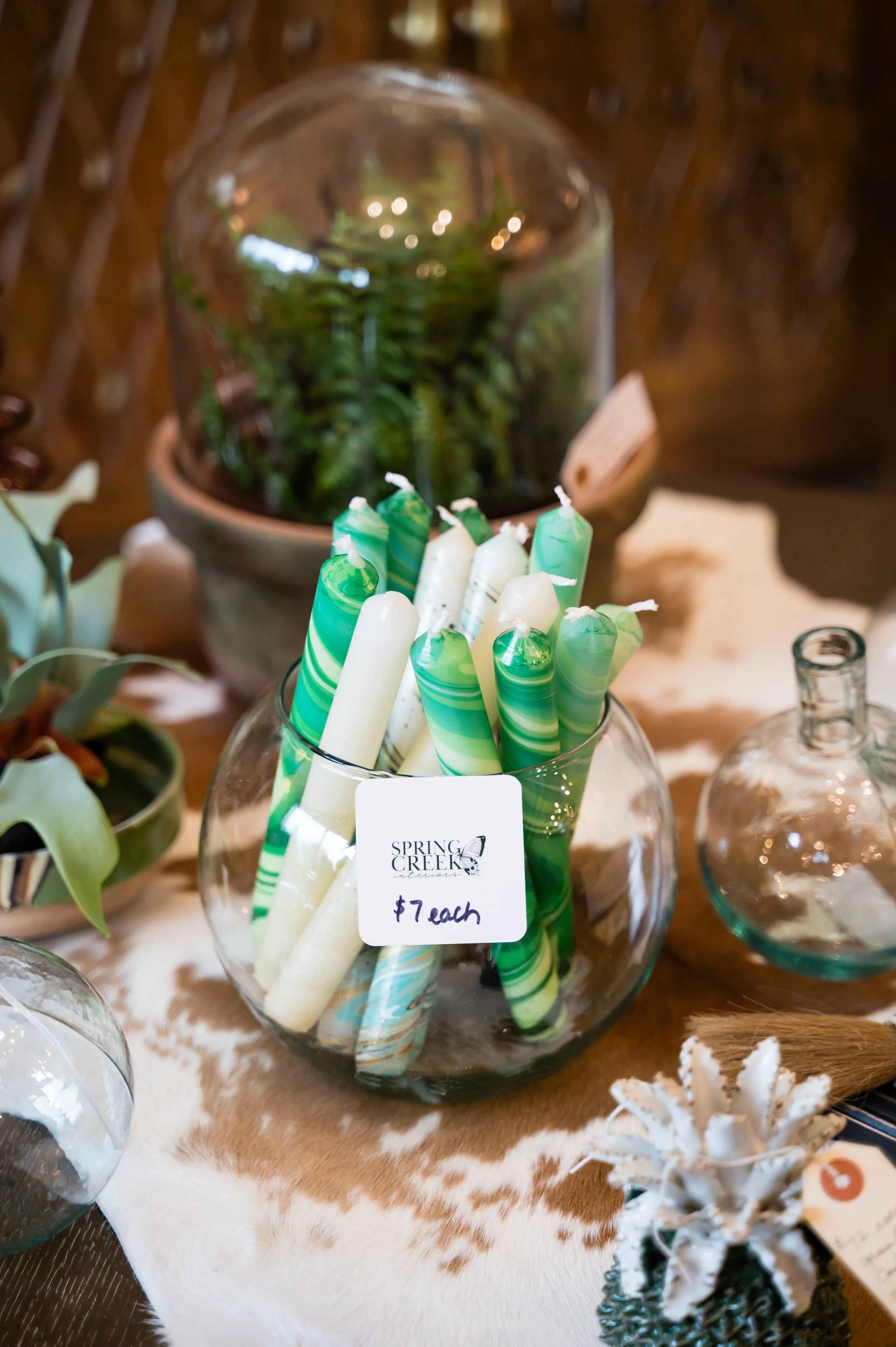 Glass bowl filled with white and green striped candles on a table with decorative items, including a potted plant in a glass terrarium and a small white coral sculpture, with a cowhide tablecloth underneath.