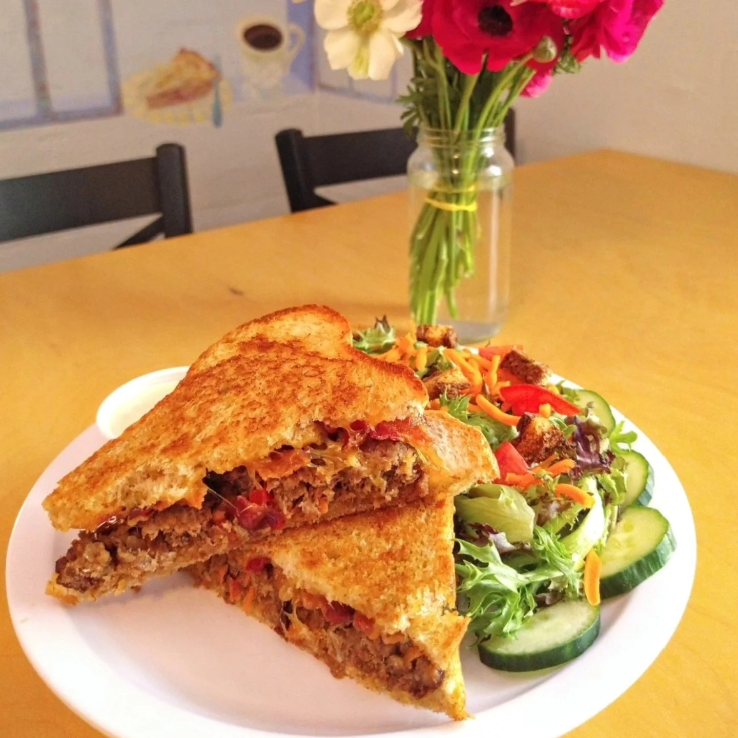 A plate with a toasted sandwich filled with ground meat and vegetables, and a side salad with lettuce, cucumber, carrots, and red peppers on a wooden table.