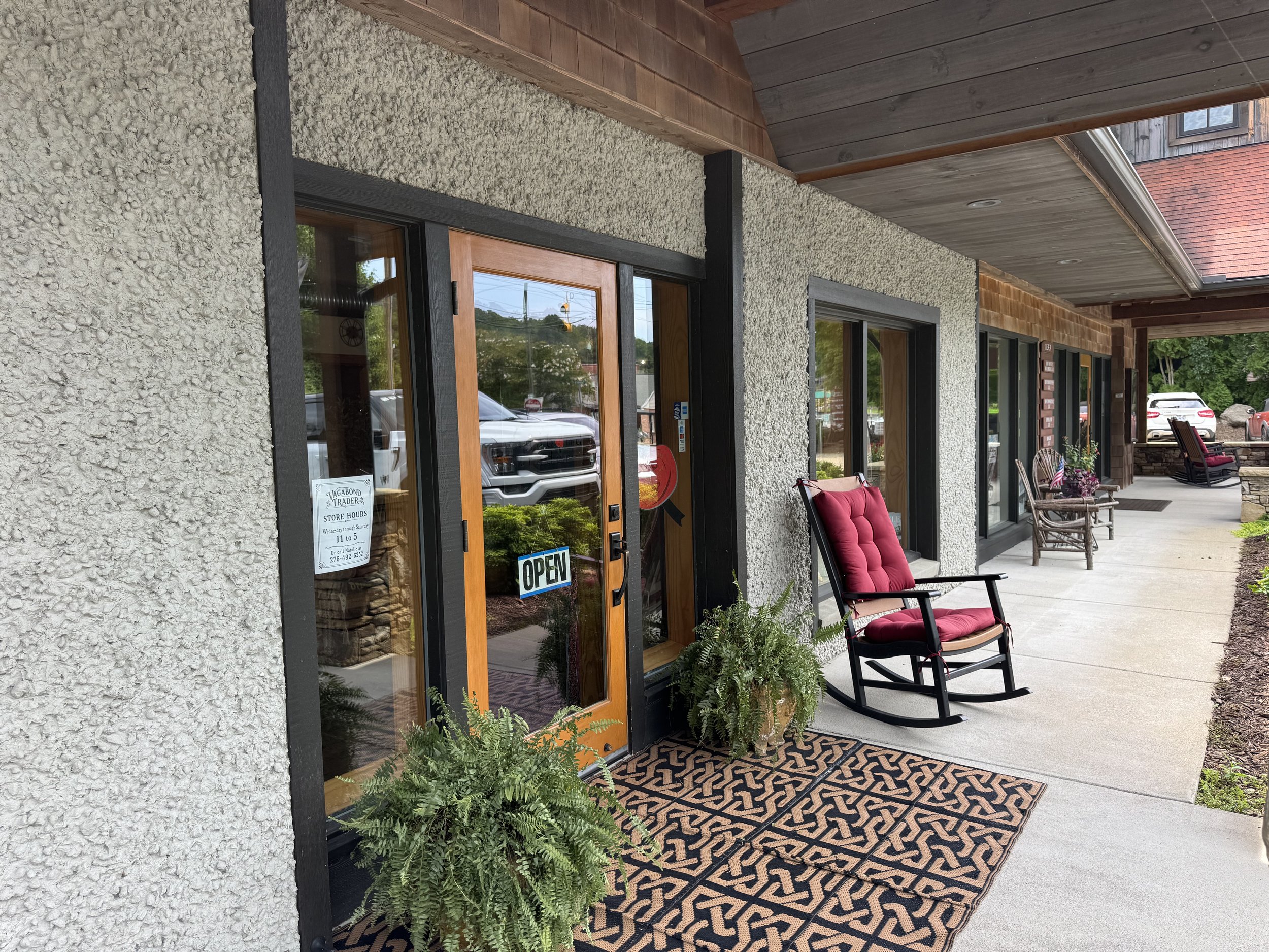 Storefront with textured stucco walls, wooden framing around front door and windows, and a covered porch area with chairs and a patterned outdoor rug. A sign in the window shows store hours, and an 'Open' sign hangs on the door. Cars are parked outside, and there's greenery in the background.