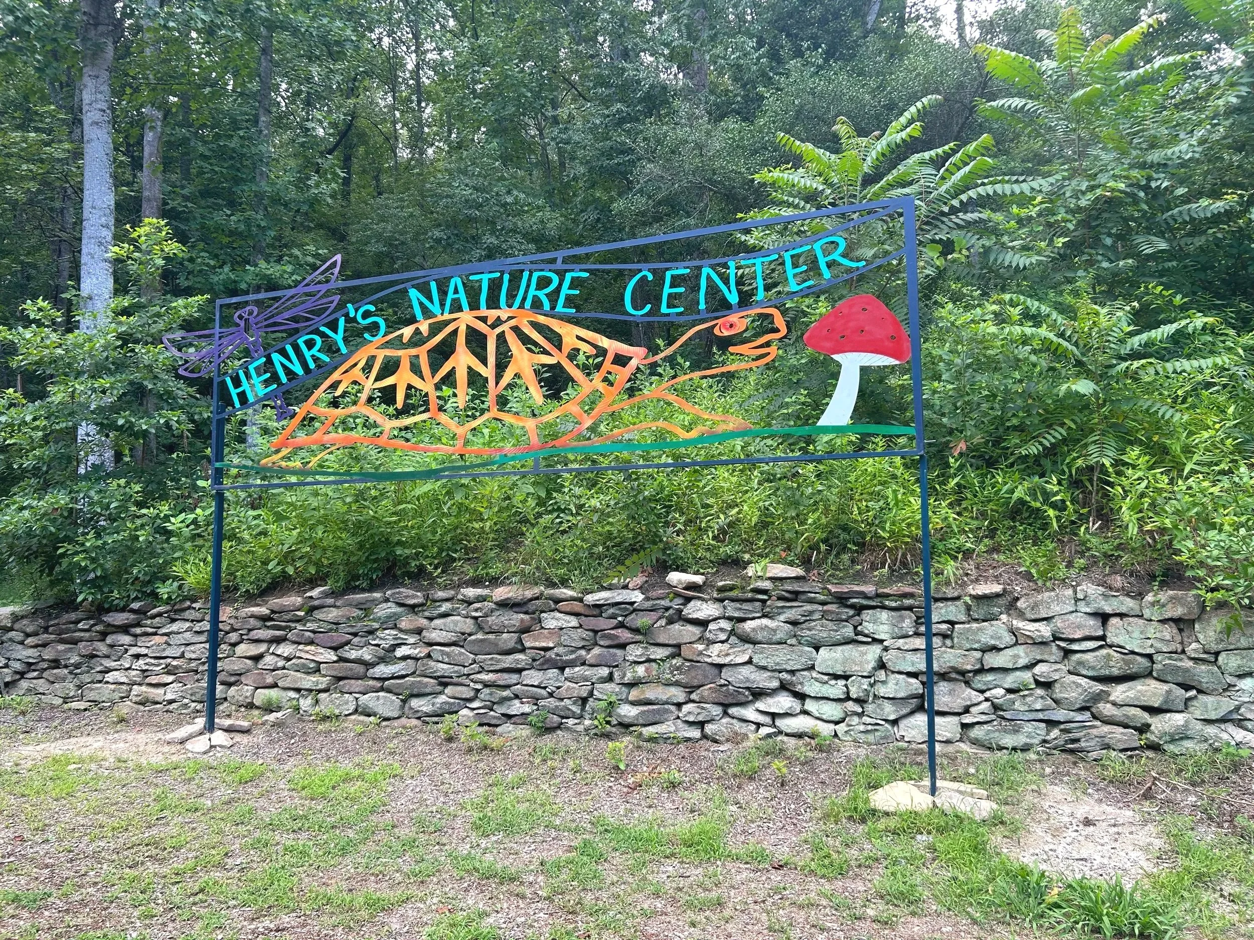 Colorful metal sign for Henry's Nature Center, featuring a dragonfly, a leaf, a mushroom, and a snake, situated in a wooded area with trees and green foliage.