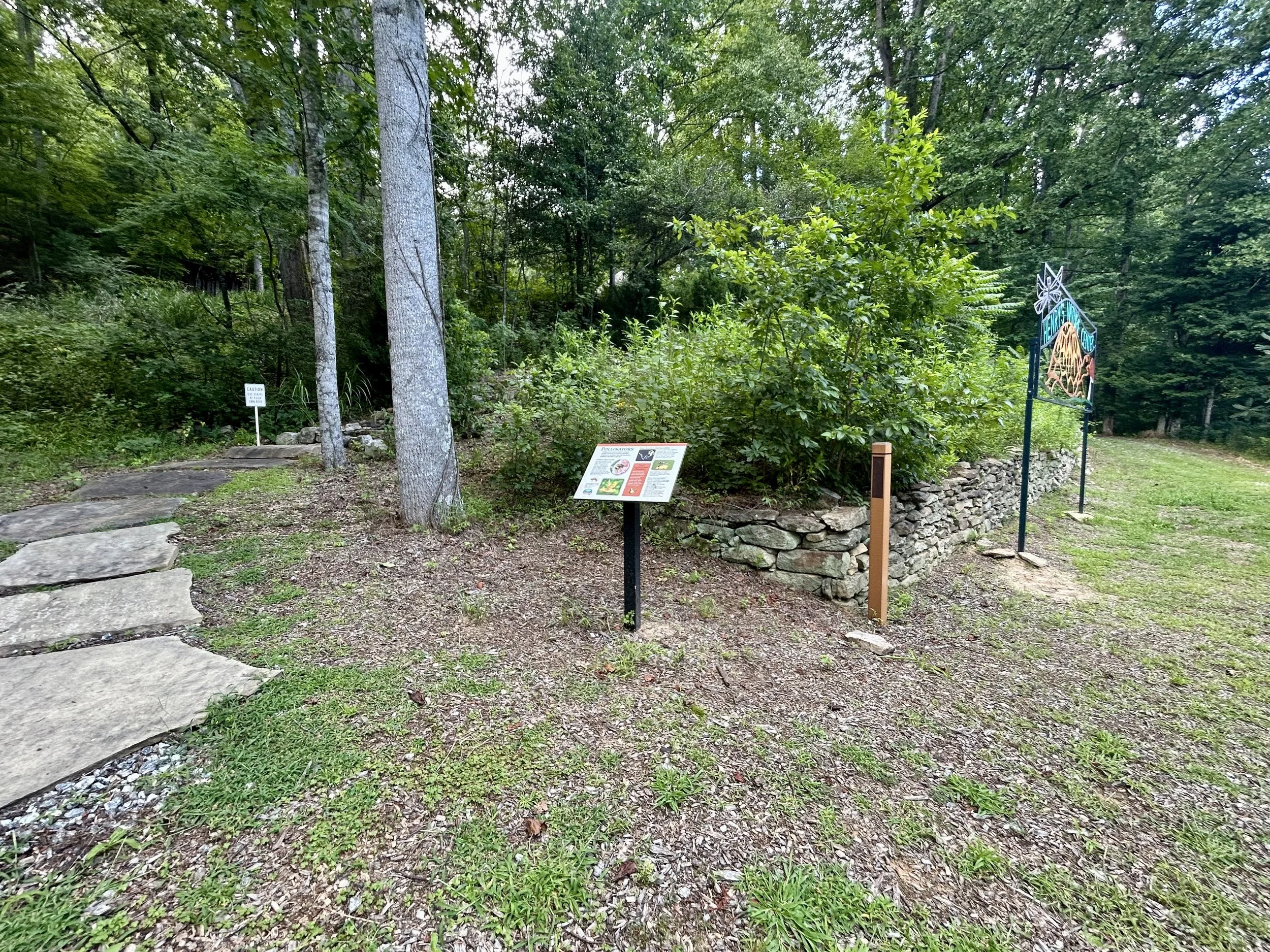 A wooded area with trees, shrubs, a stone path in the foreground, a sign, and a decorative metal art piece on the right side.