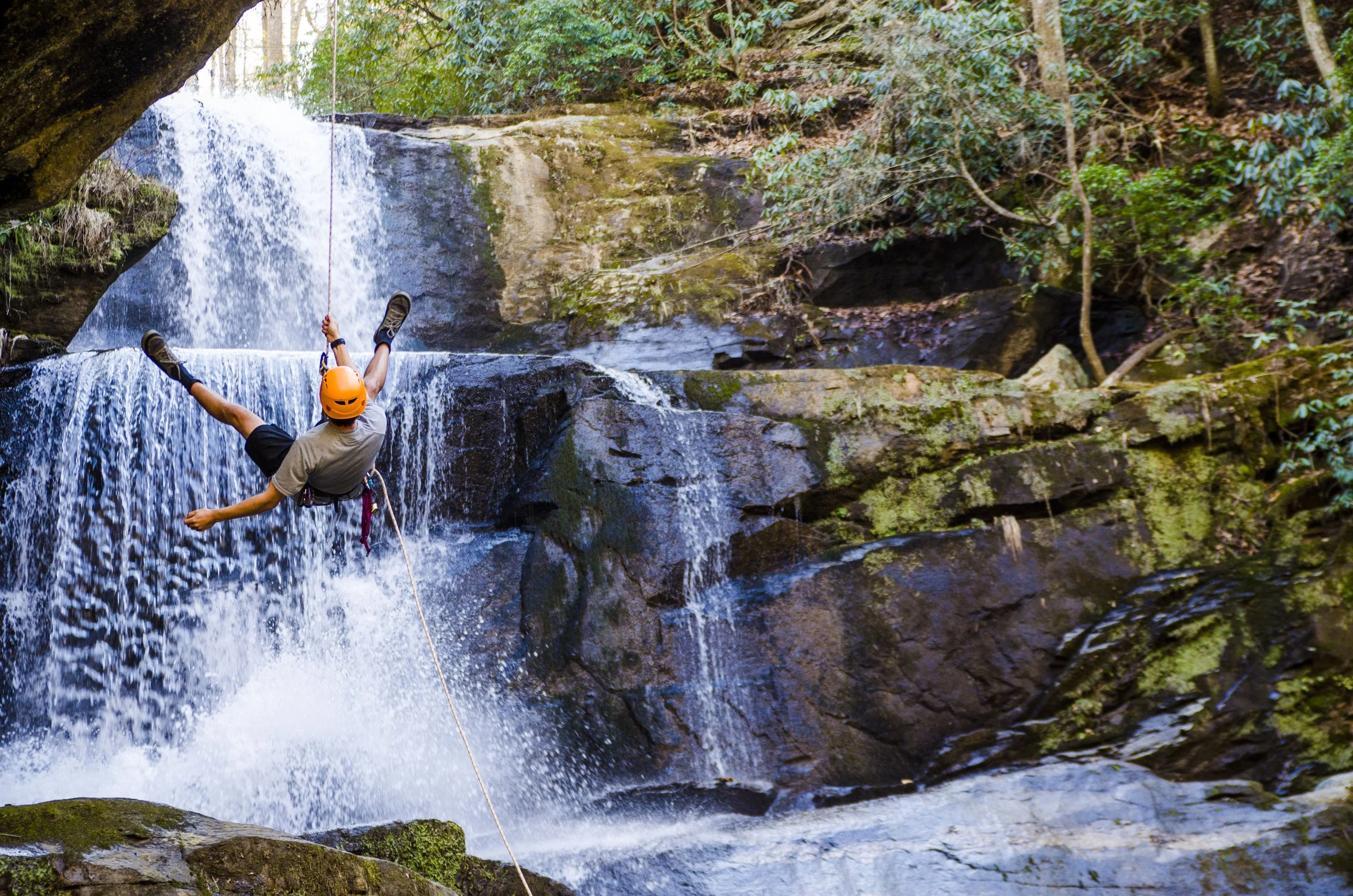 A person wearing a helmet and outdoor gear rappelling down a small waterfall in a forested area.
