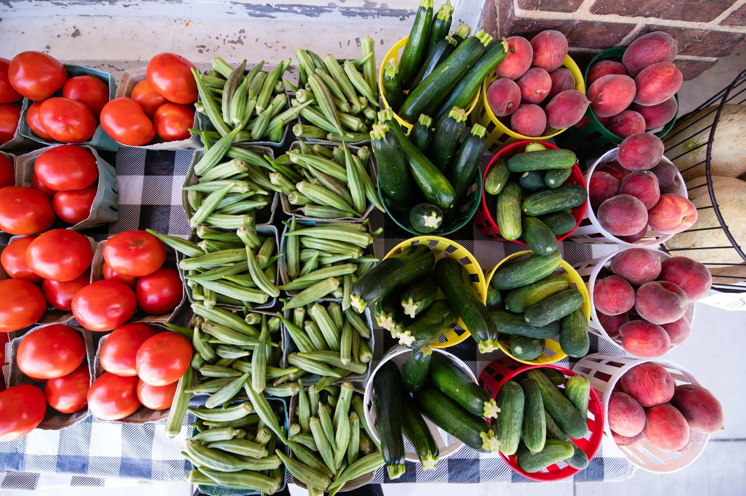 Various fresh vegetables displayed in baskets and containers on a table, including tomatoes, okra, zucchini, peaches, and cantaloupe.