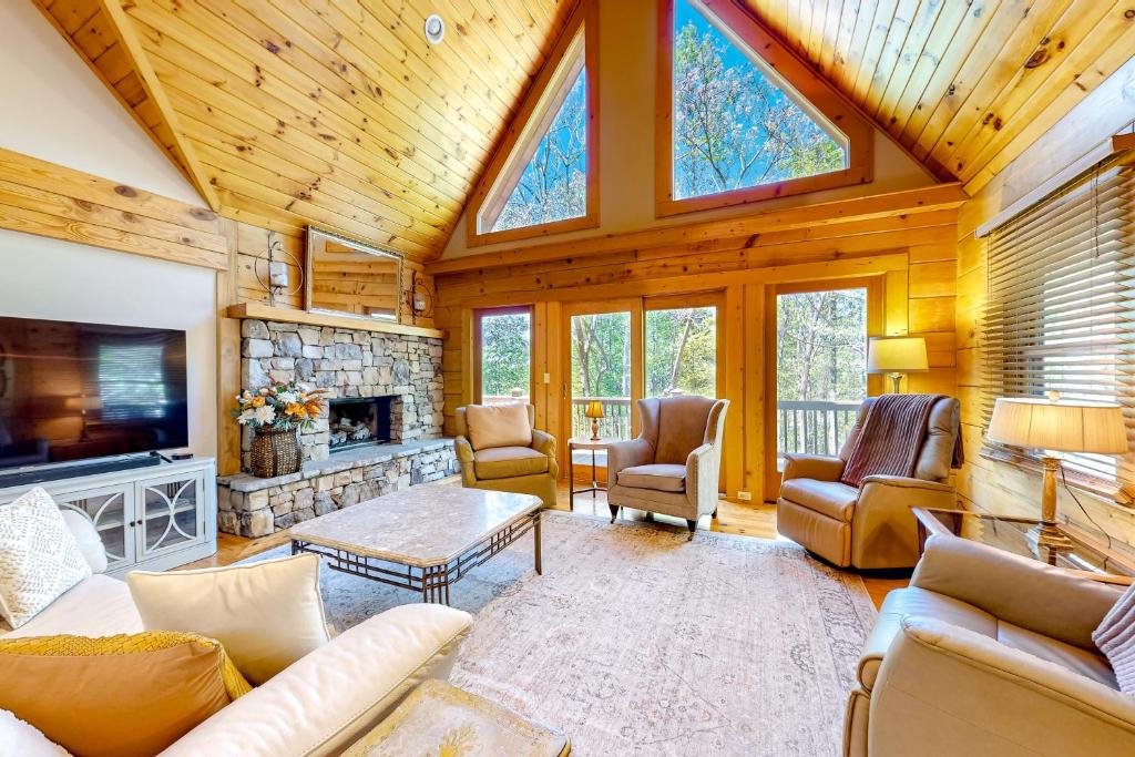 Cozy living room with wood-paneled walls and ceiling, large windows with a forest view, stone fireplace, and comfortable seating.