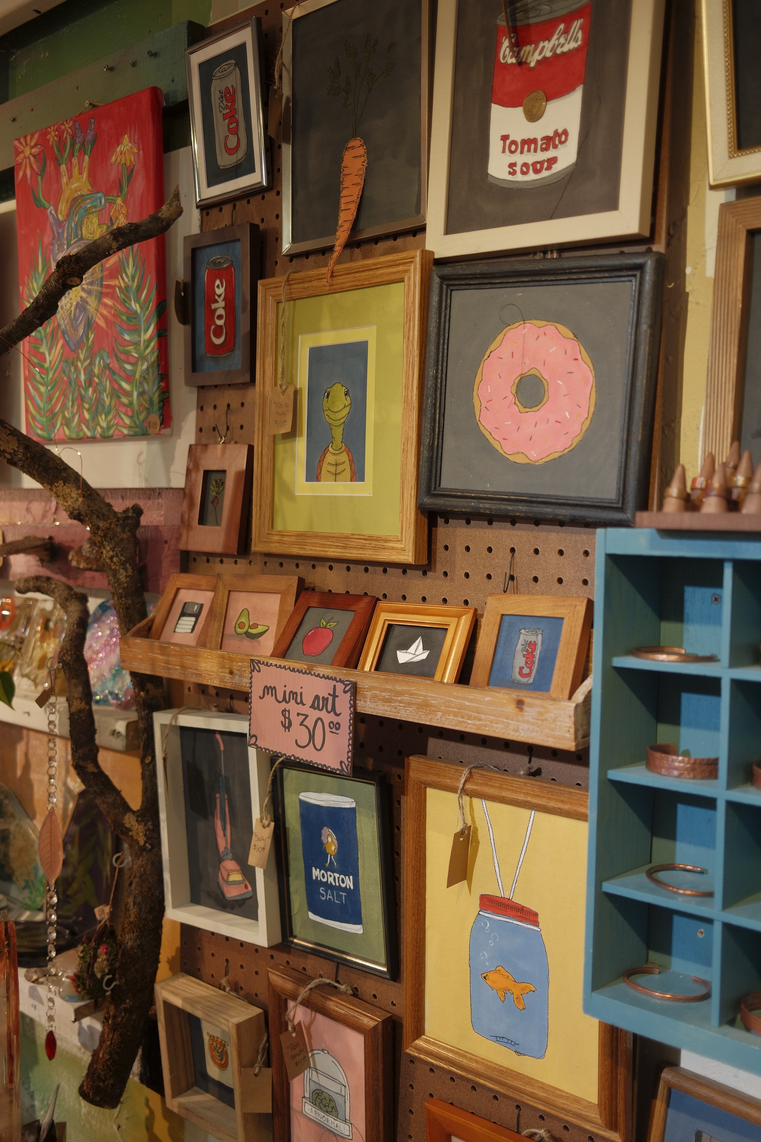 Wall of colorful paintings including a carrot, Coca-Cola cans, a donut, a fish in a jar, and other framed art in various styles and sizes.