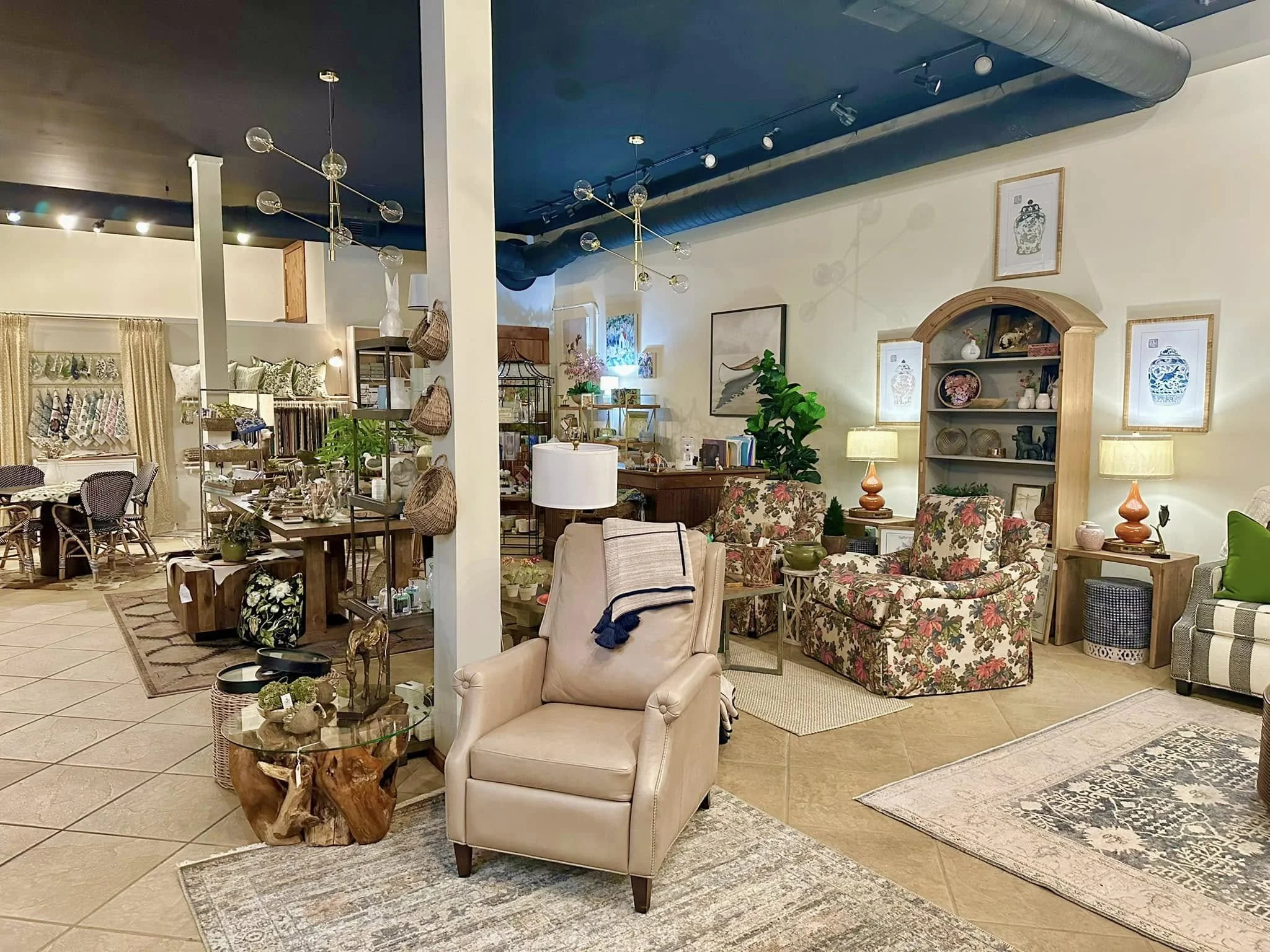 Interior of a furniture store with various chairs, tables, and decorative items arranged in a room with patterned rugs, framed artwork on the walls, and shelving displaying vases and sculptures.