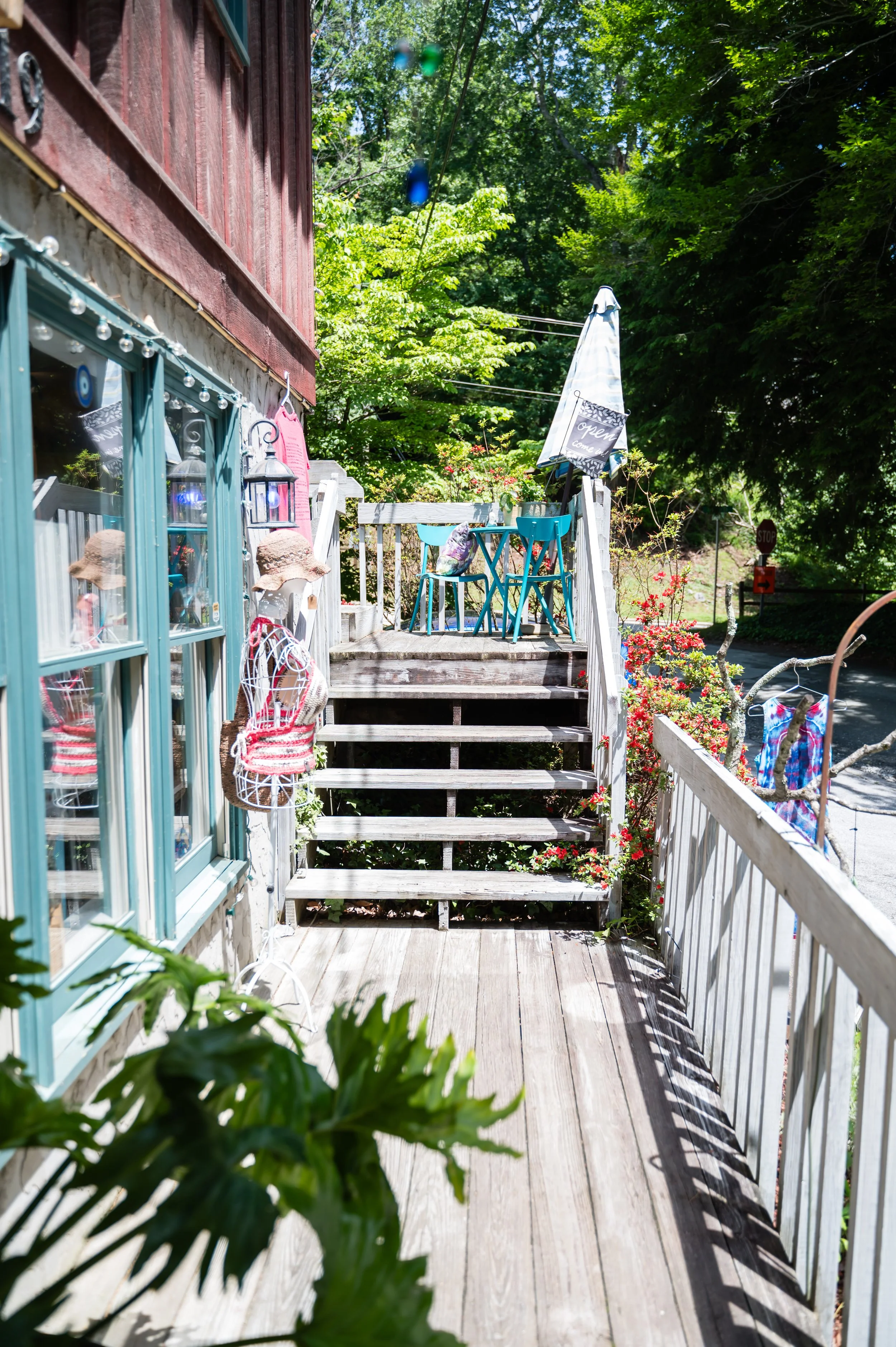 A small outdoor wooden deck with a turquoise table and two turquoise chairs, decorated with potted plants and a large umbrella, surrounded by lush green trees and a white railing.